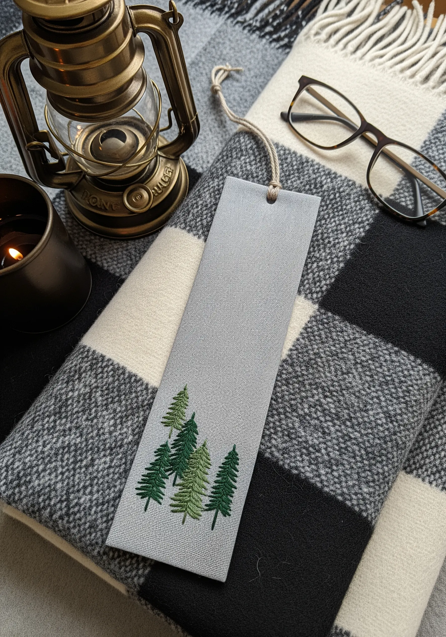 Embroidered bookmark with a cluster of pine trees in varying shades of green on gray fabric.