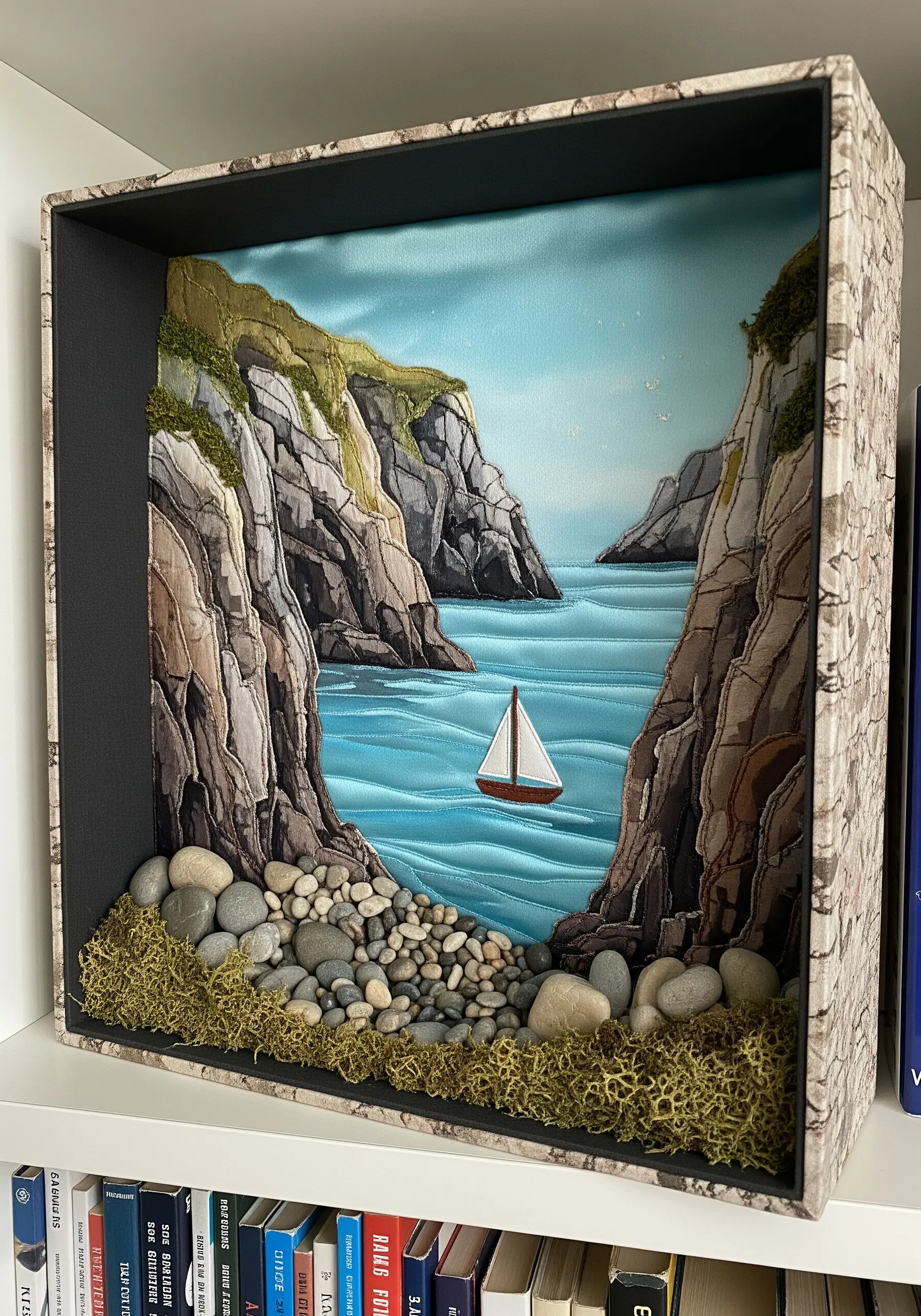 A shadow box diorama of a coastal cove with fabric cliffs, a stitched sea, and real pebbles.