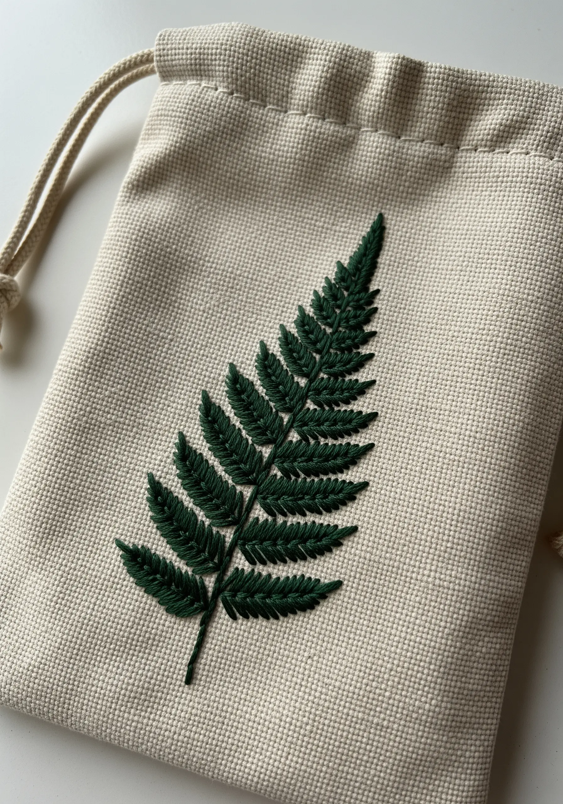 A detailed, dark green fern leaf embroidered on a small canvas drawstring bag.