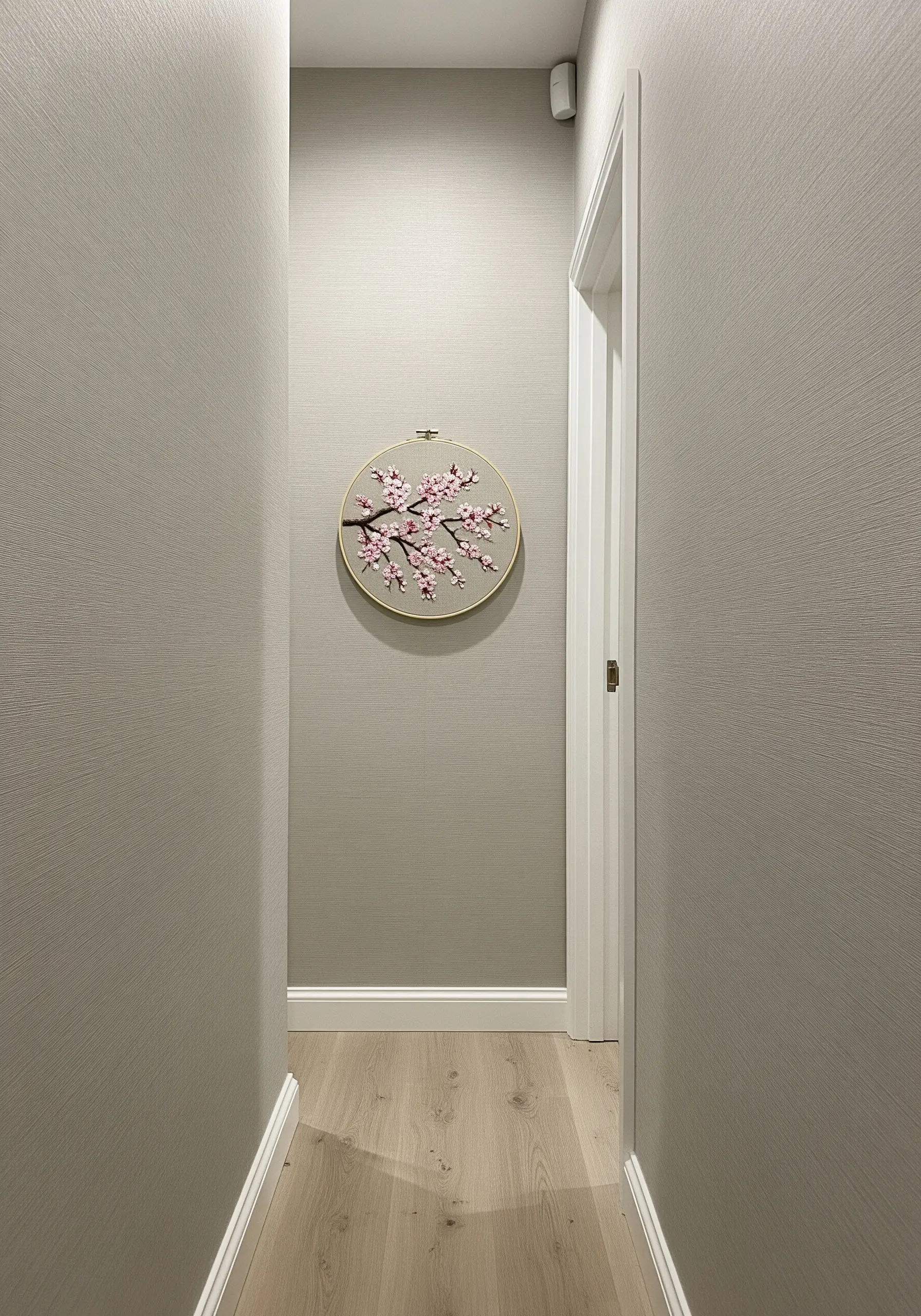 A single embroidery hoop with a cherry blossom branch hanging in a narrow, neutral-toned hallway.