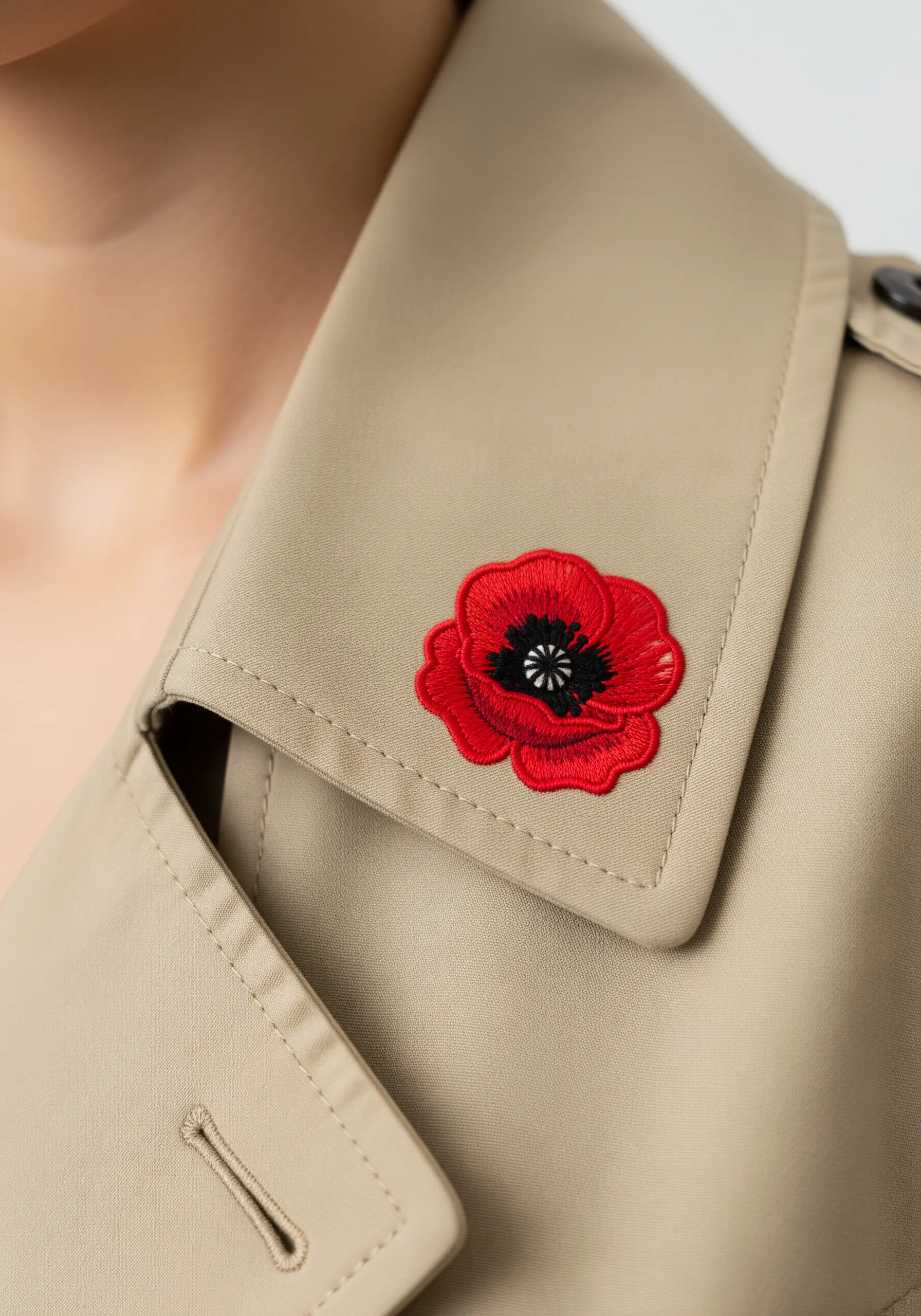 A vibrant red and black poppy flower embroidered on the lapel of a beige trench coat.