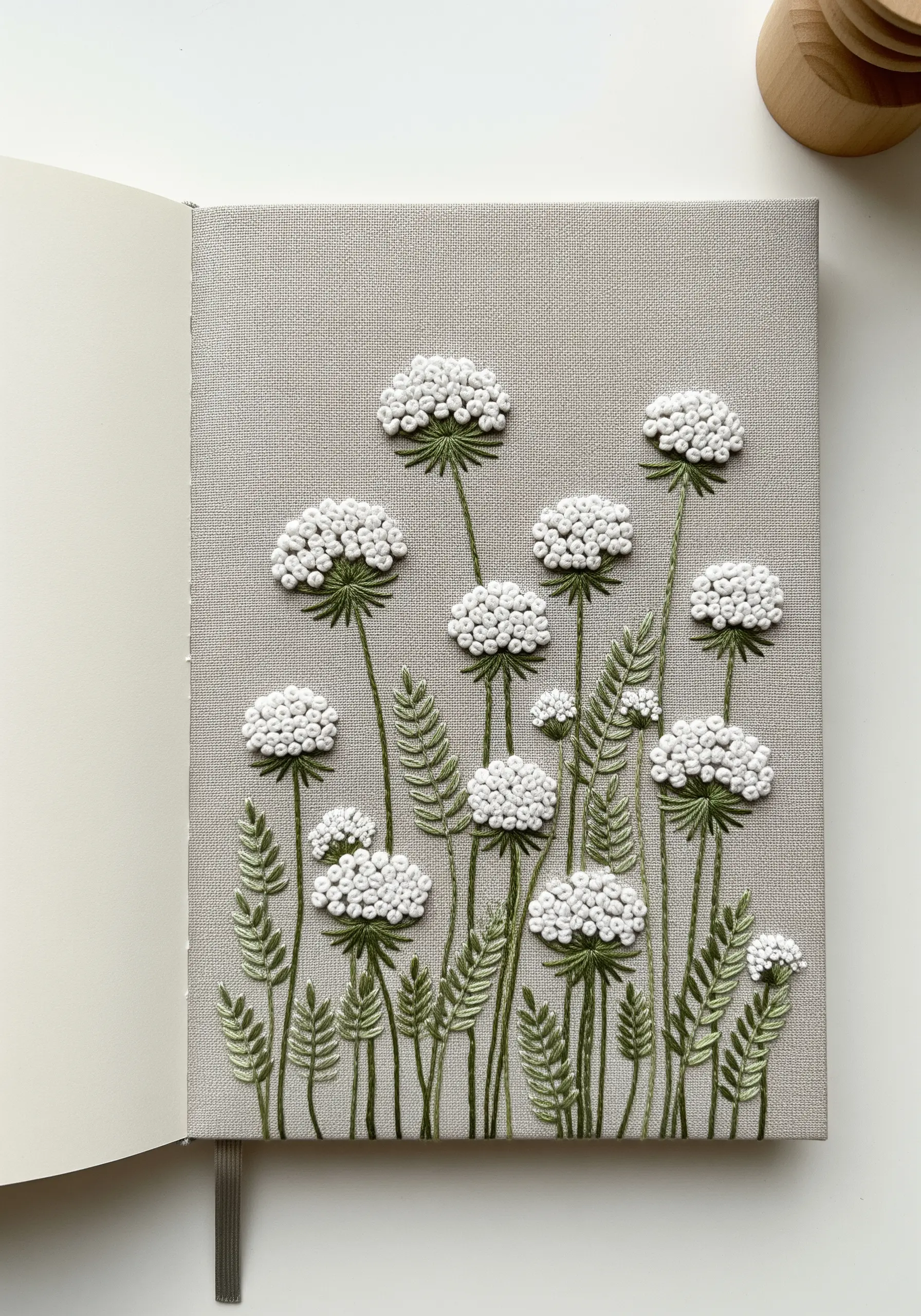 Dense white French knots creating Queen Anne's Lace flowers on a grey fabric-covered notebook.