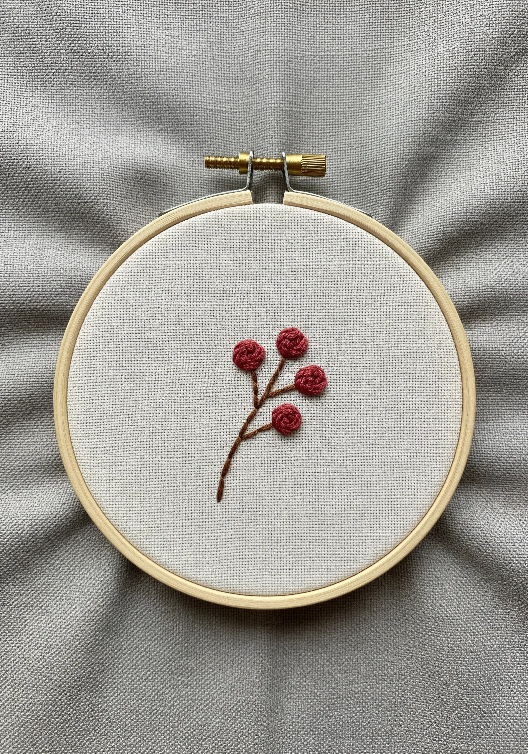 A small branch with four red, textured woven wheel stitch roses in an embroidery hoop.