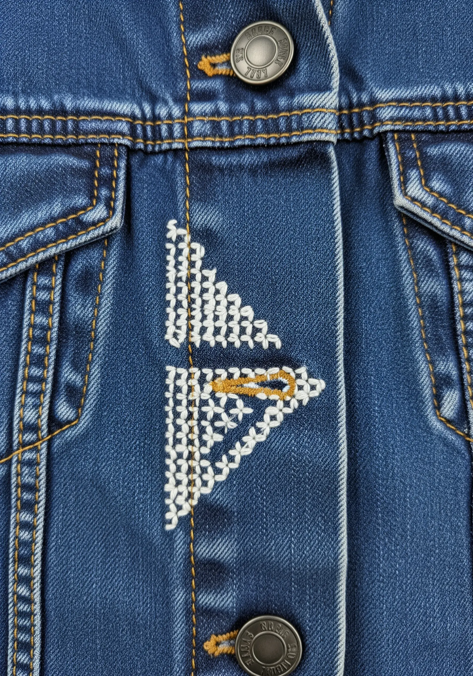 A geometric arrow motif embroidered in white and gold thread next to jacket buttons.