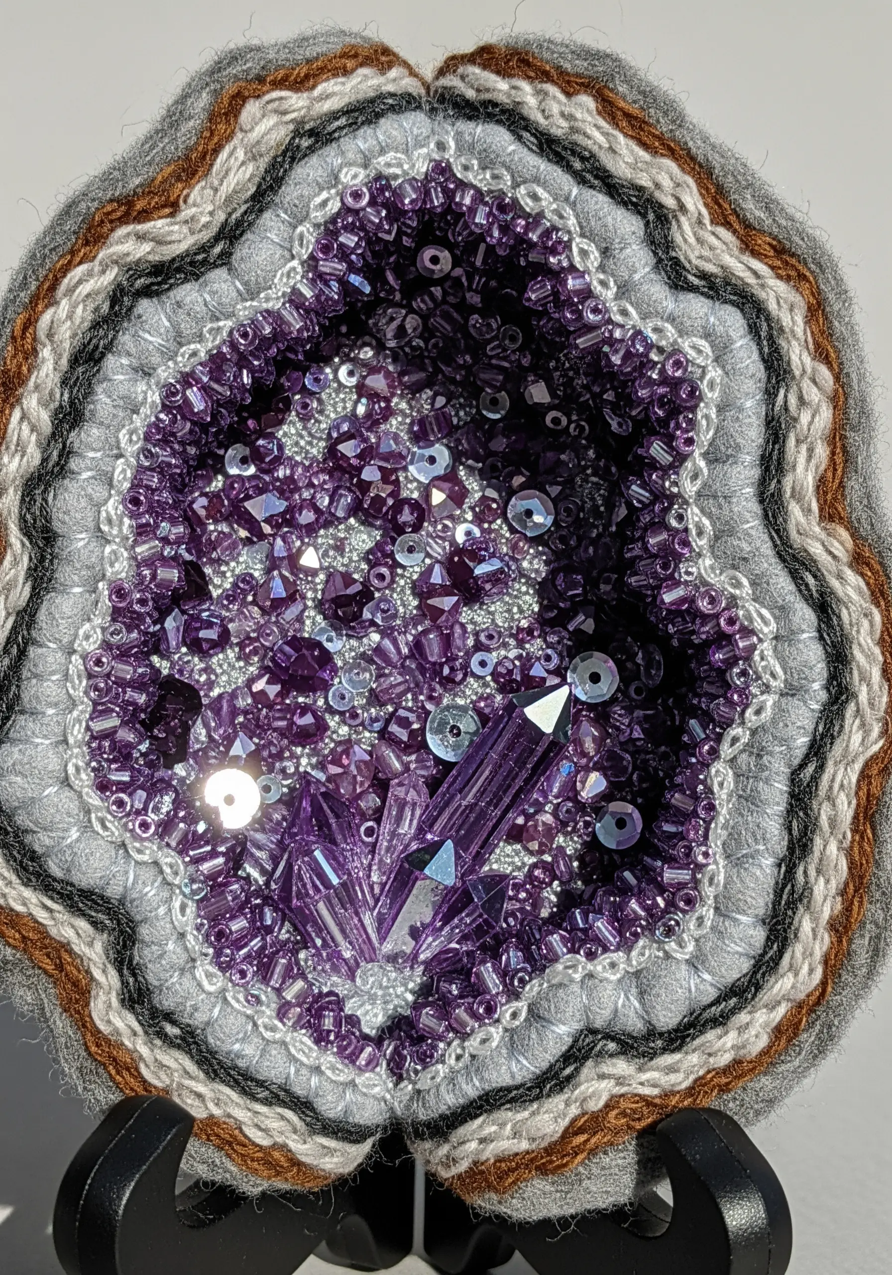 A fiber art geode made with felt, yarn, and a dense cluster of purple beads and crystals.