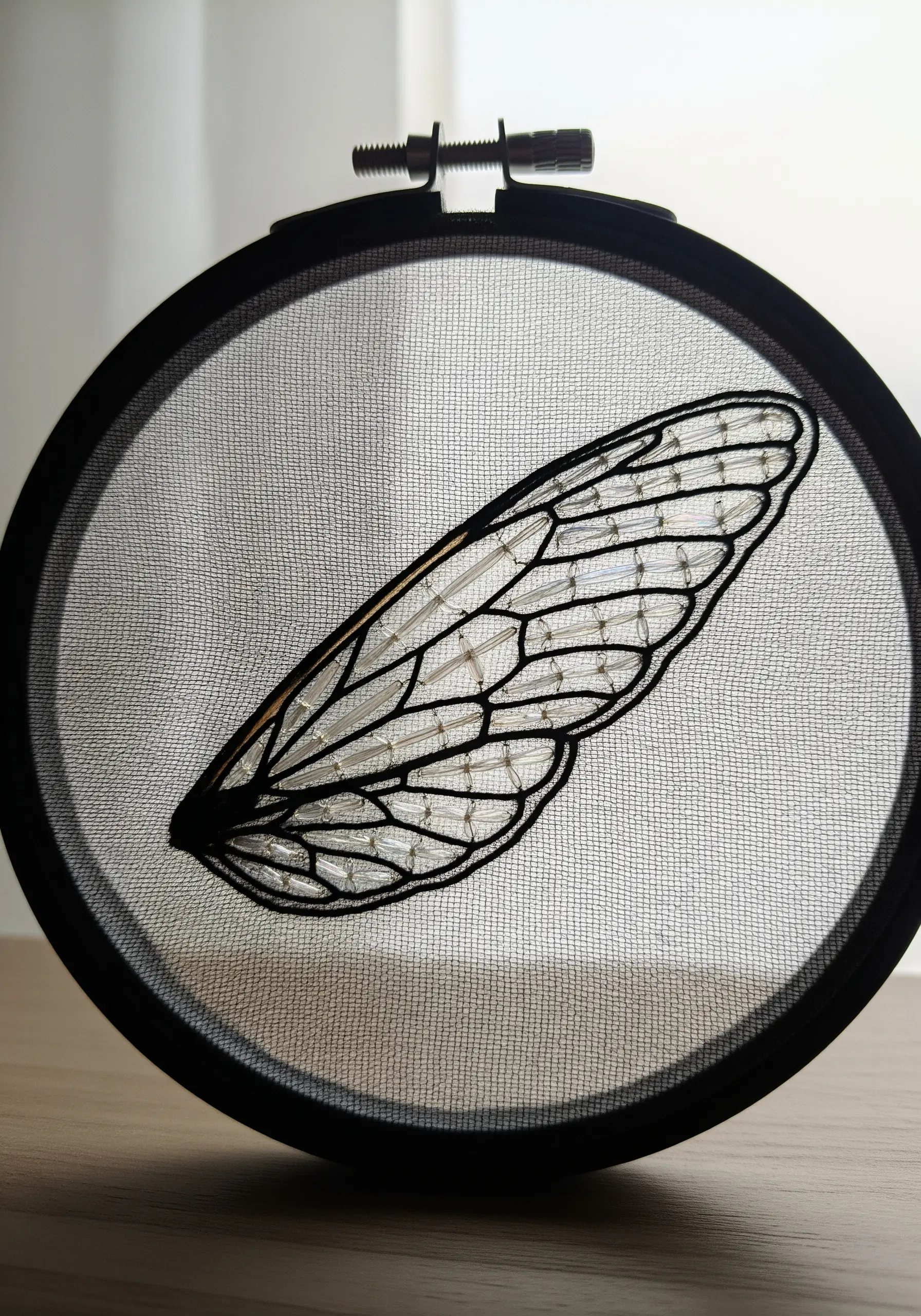 A delicate insect wing embroidered in black thread on see-through fabric in a black hoop.