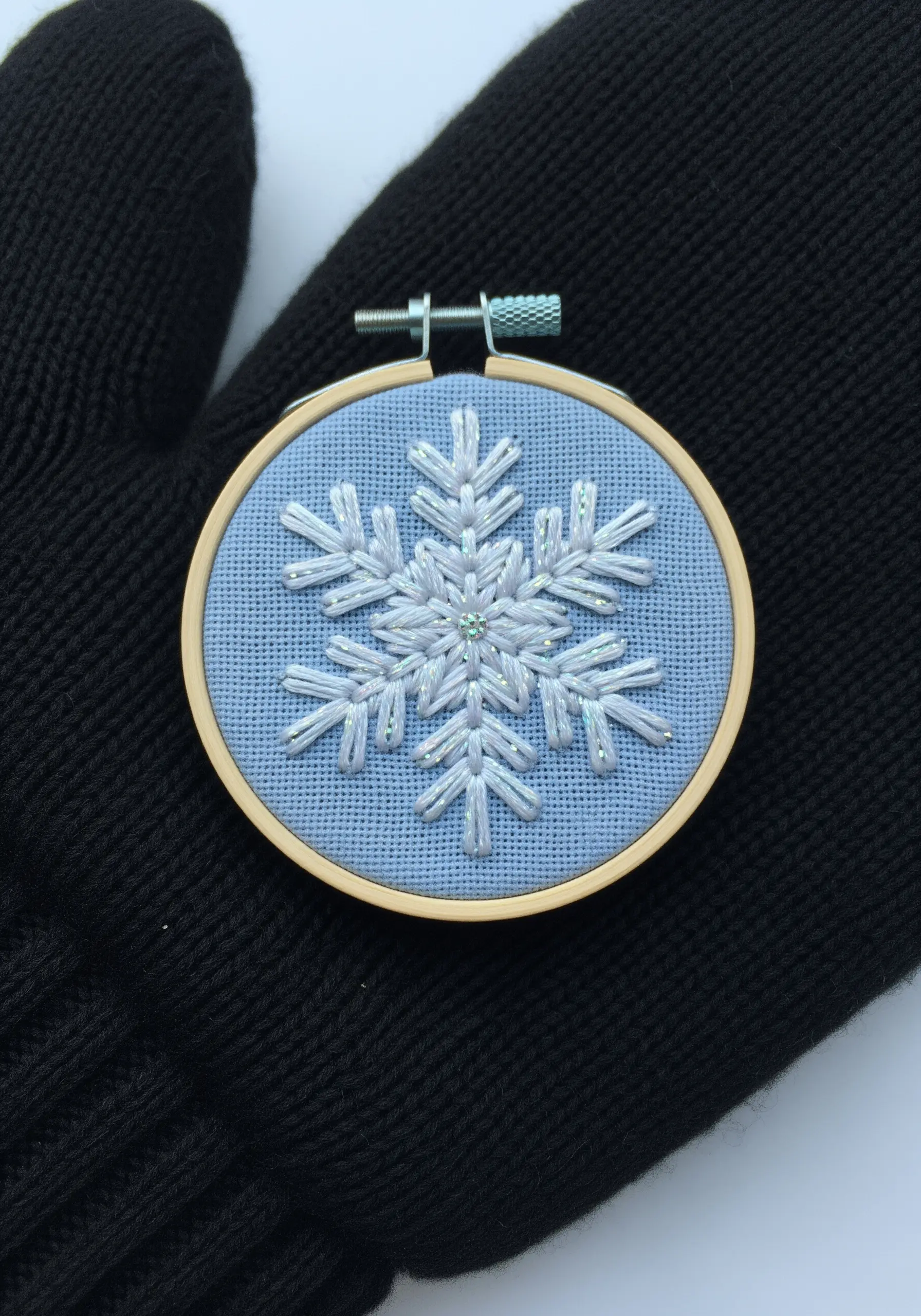 A white and iridescent embroidered snowflake on a blue background, held by a black glove.