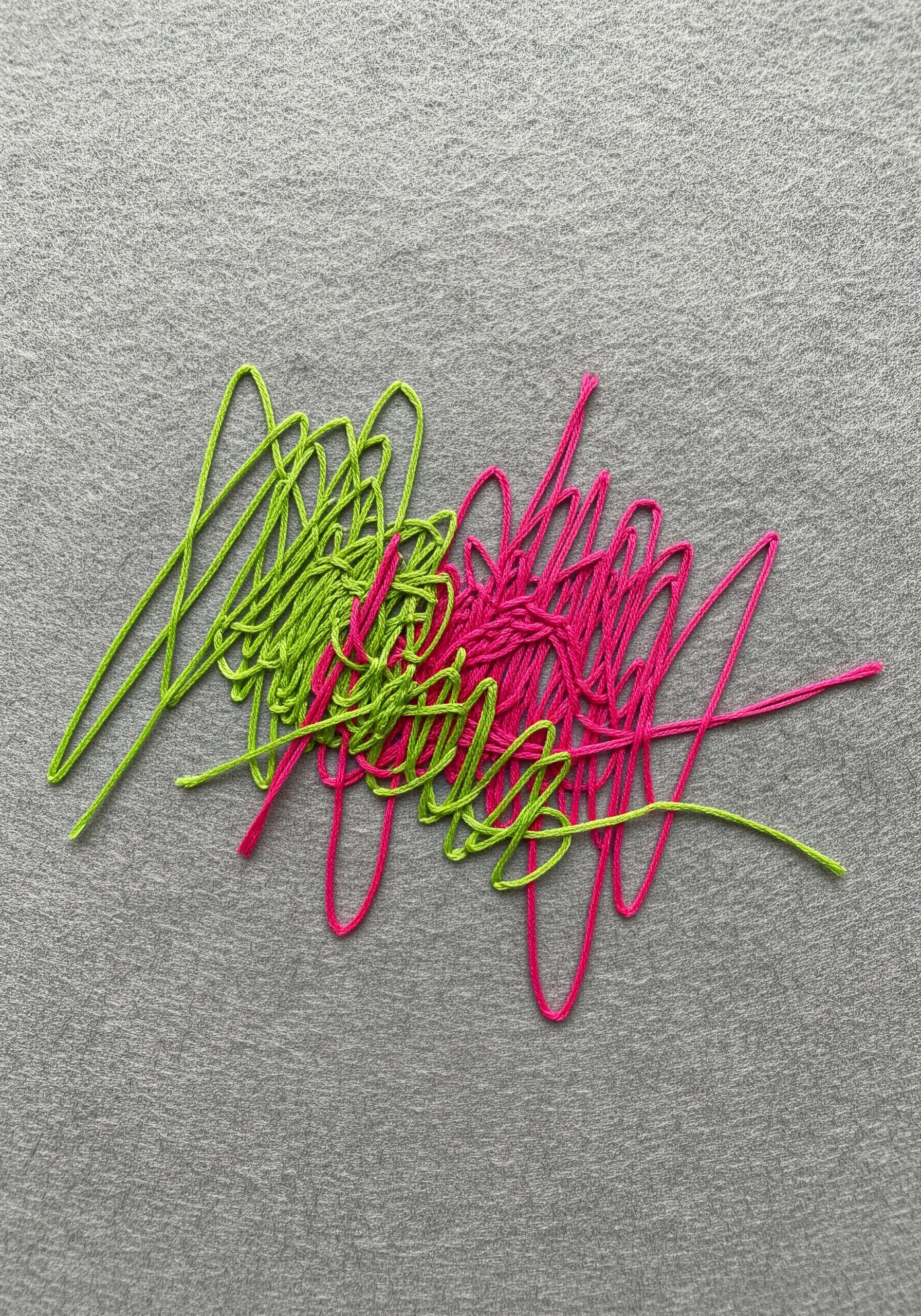 An abstract embroidery of intersecting green and pink scribbled thread on gray fabric.