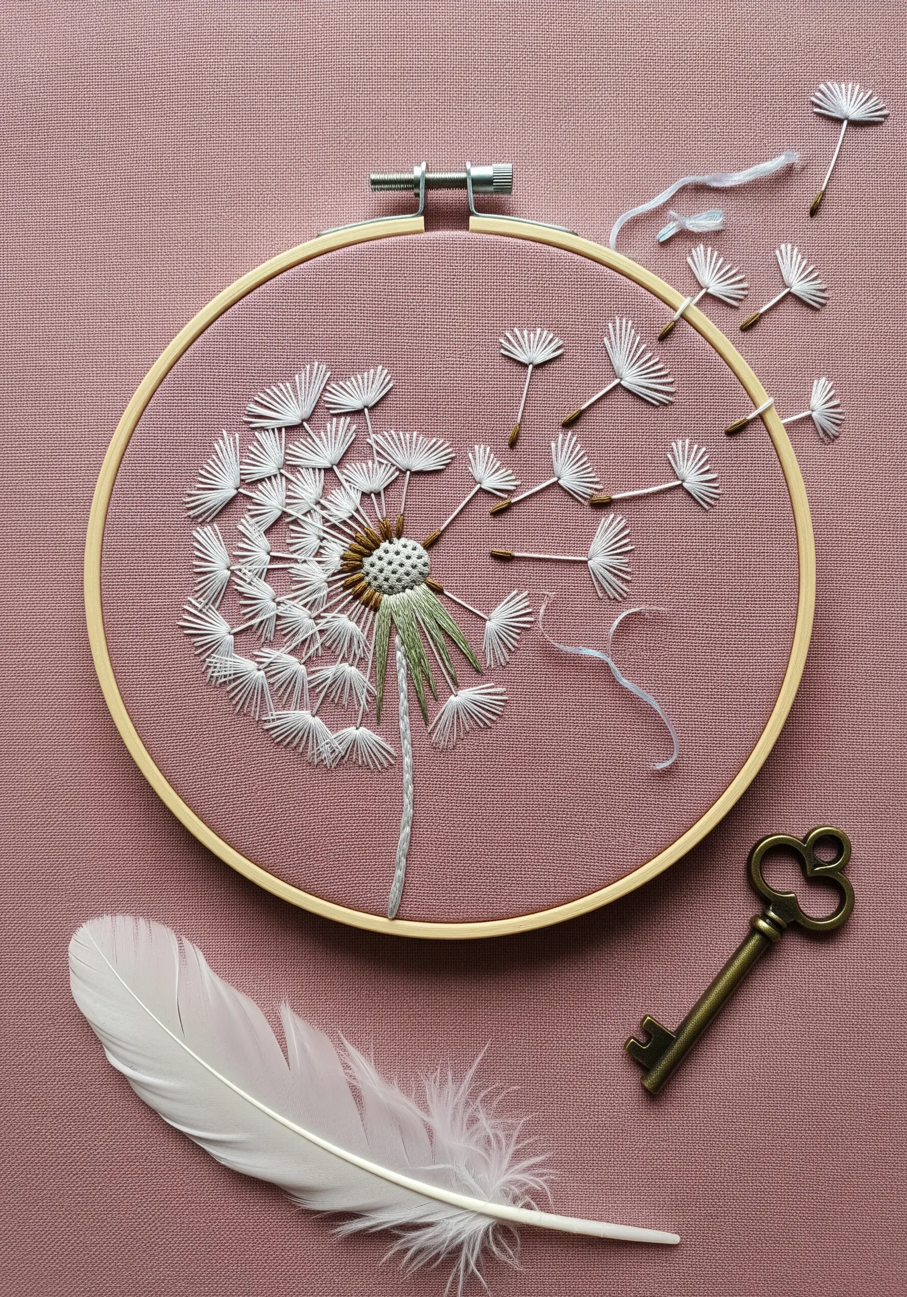 A dandelion puff with seeds blowing away, stitched in white on a dusty pink fabric.