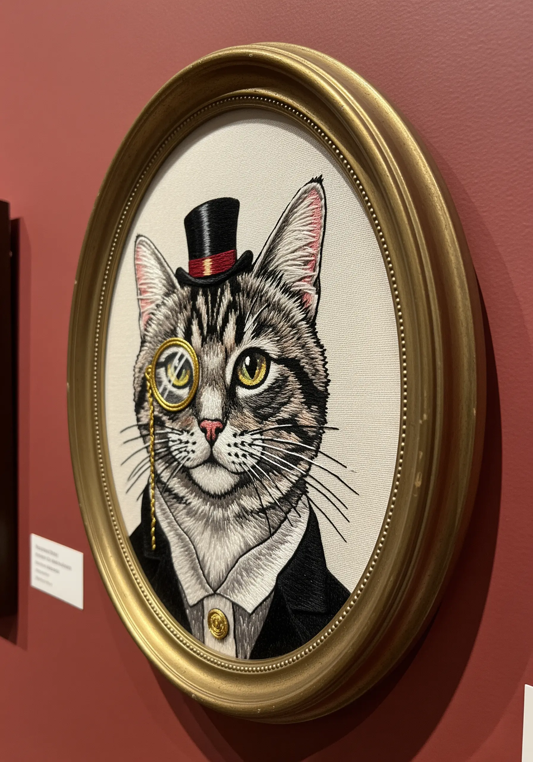 A highly detailed embroidered portrait of a tabby cat wearing a top hat and monocle, in a gold frame.