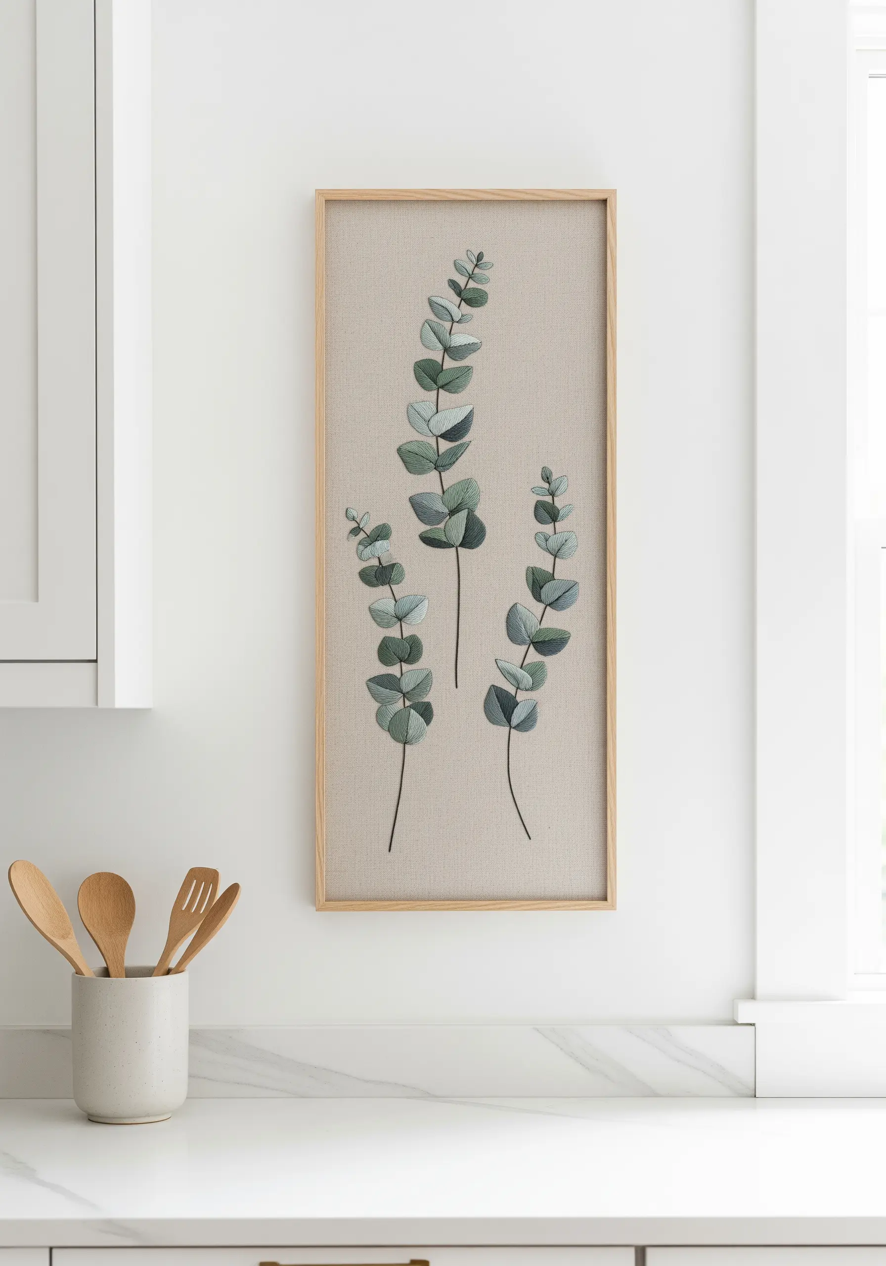 A framed rectangular embroidery of three eucalyptus stems with muted green leaves.