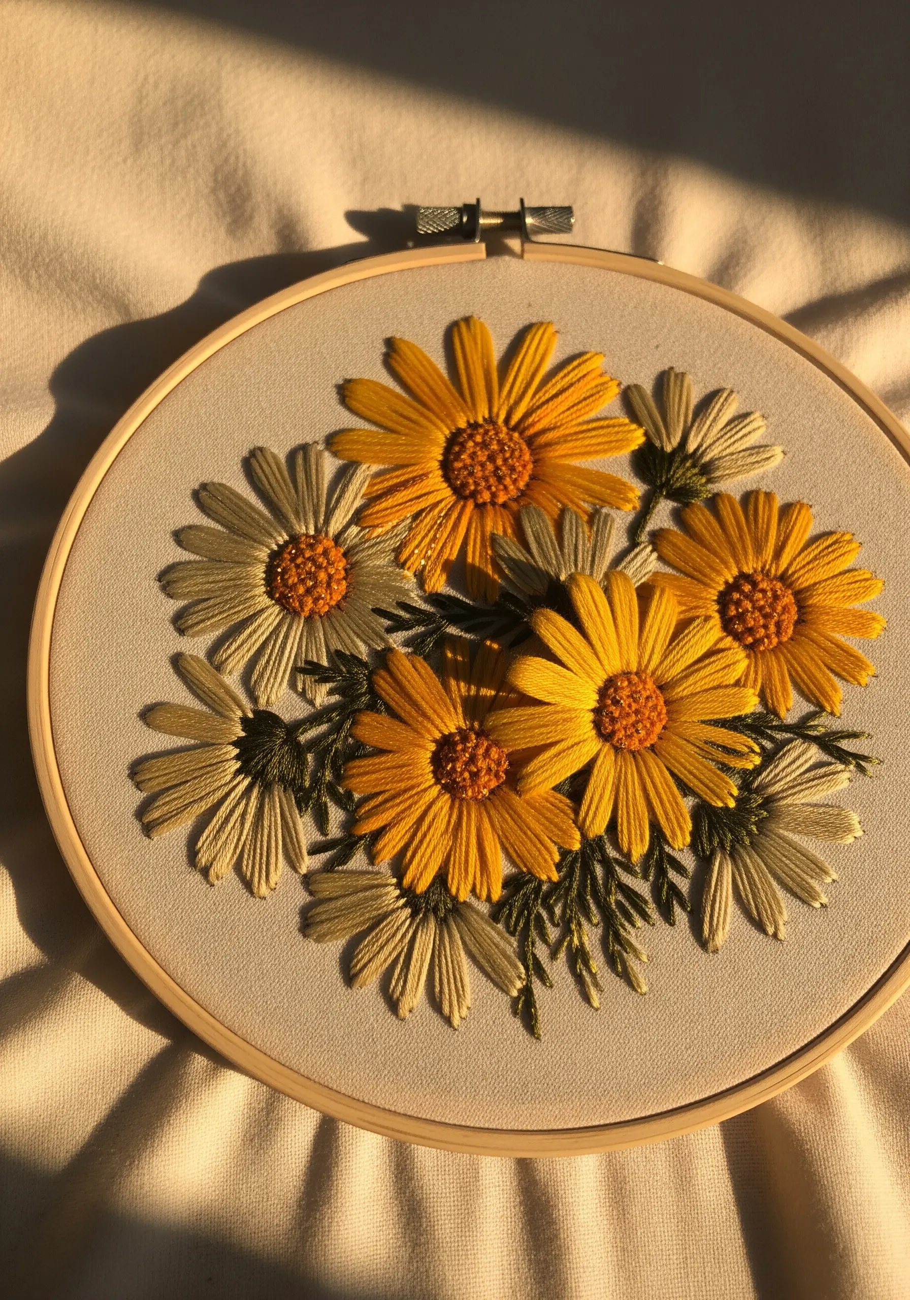 A bouquet of golden-yellow and muted-white daisies embroidered in a hoop, lit by warm sunlight.