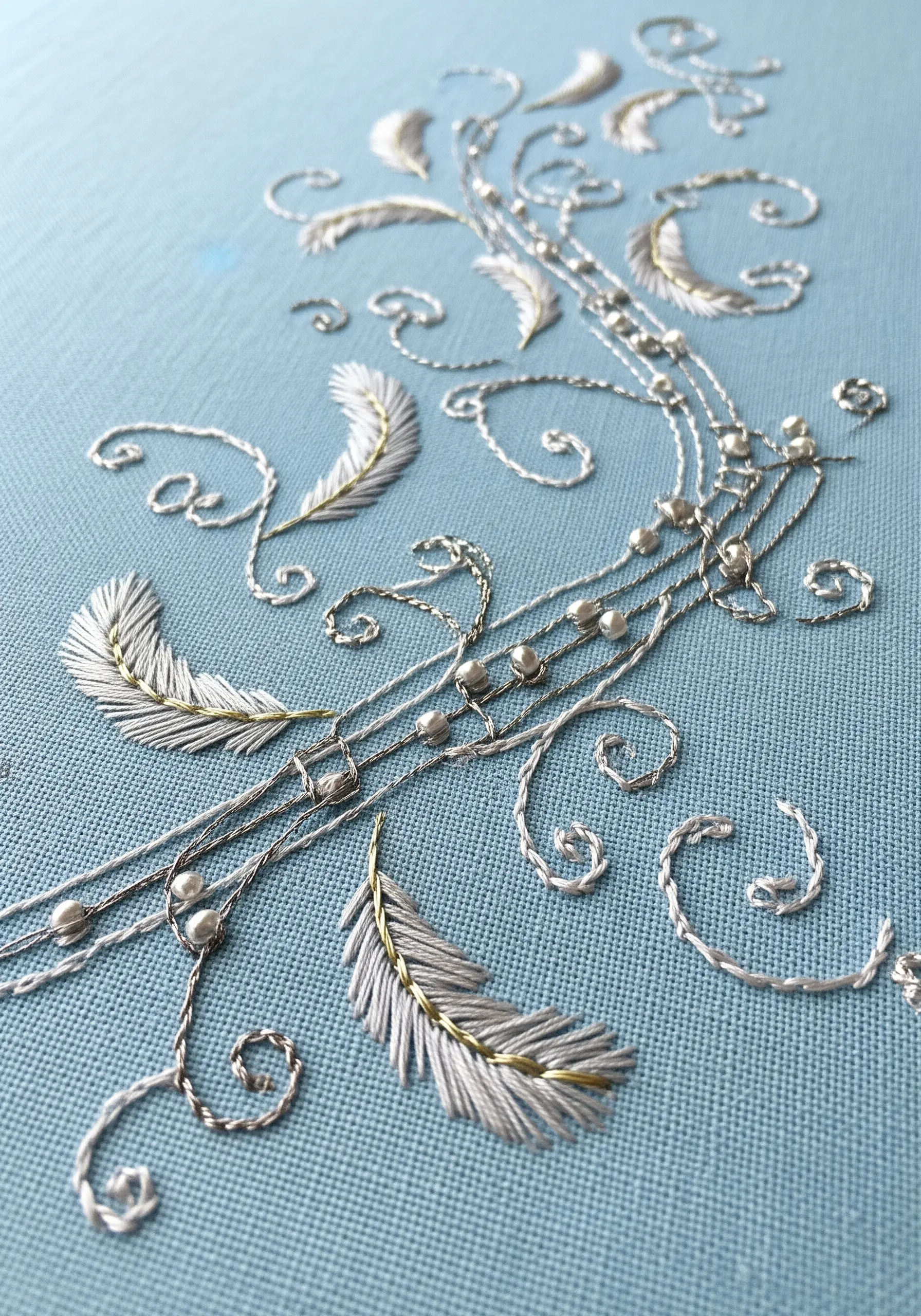 Delicate embroidery with silver and gold metallic threads, beads, and feather motifs on blue fabric.