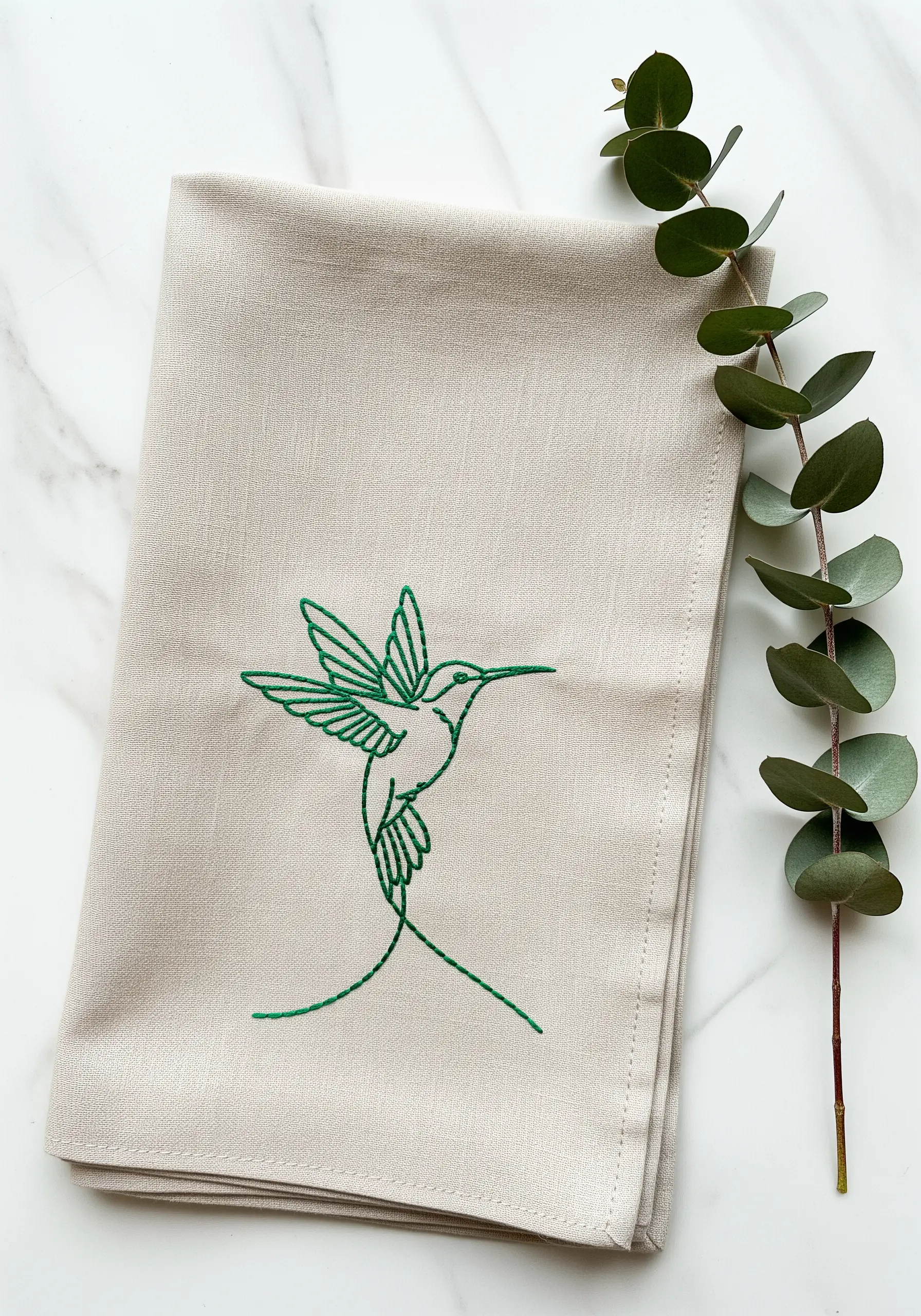 Emerald green line art hummingbird embroidered on a natural linen napkin.