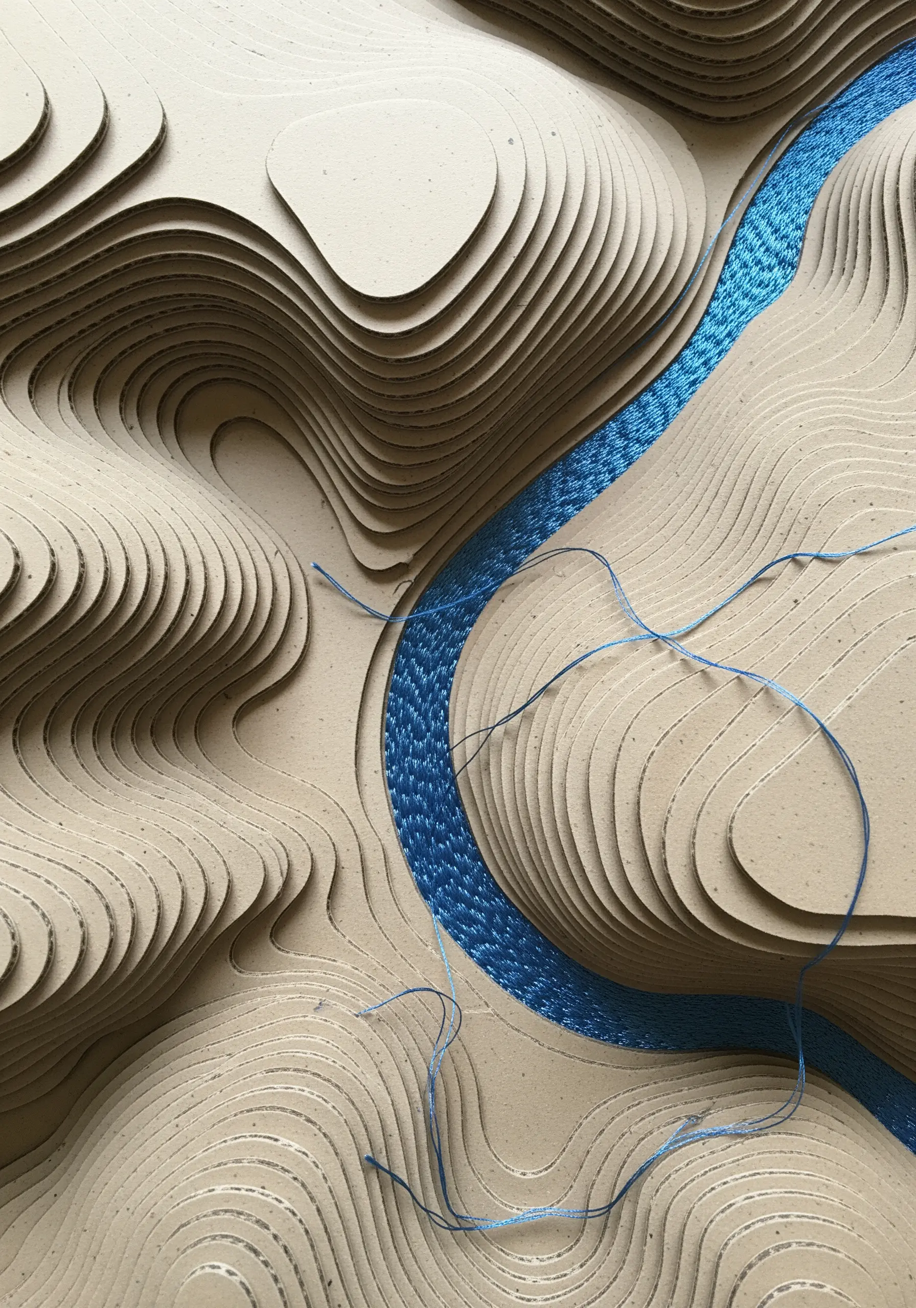 A topographical map made from layered cardboard, with a shimmering blue thread couched as a river.