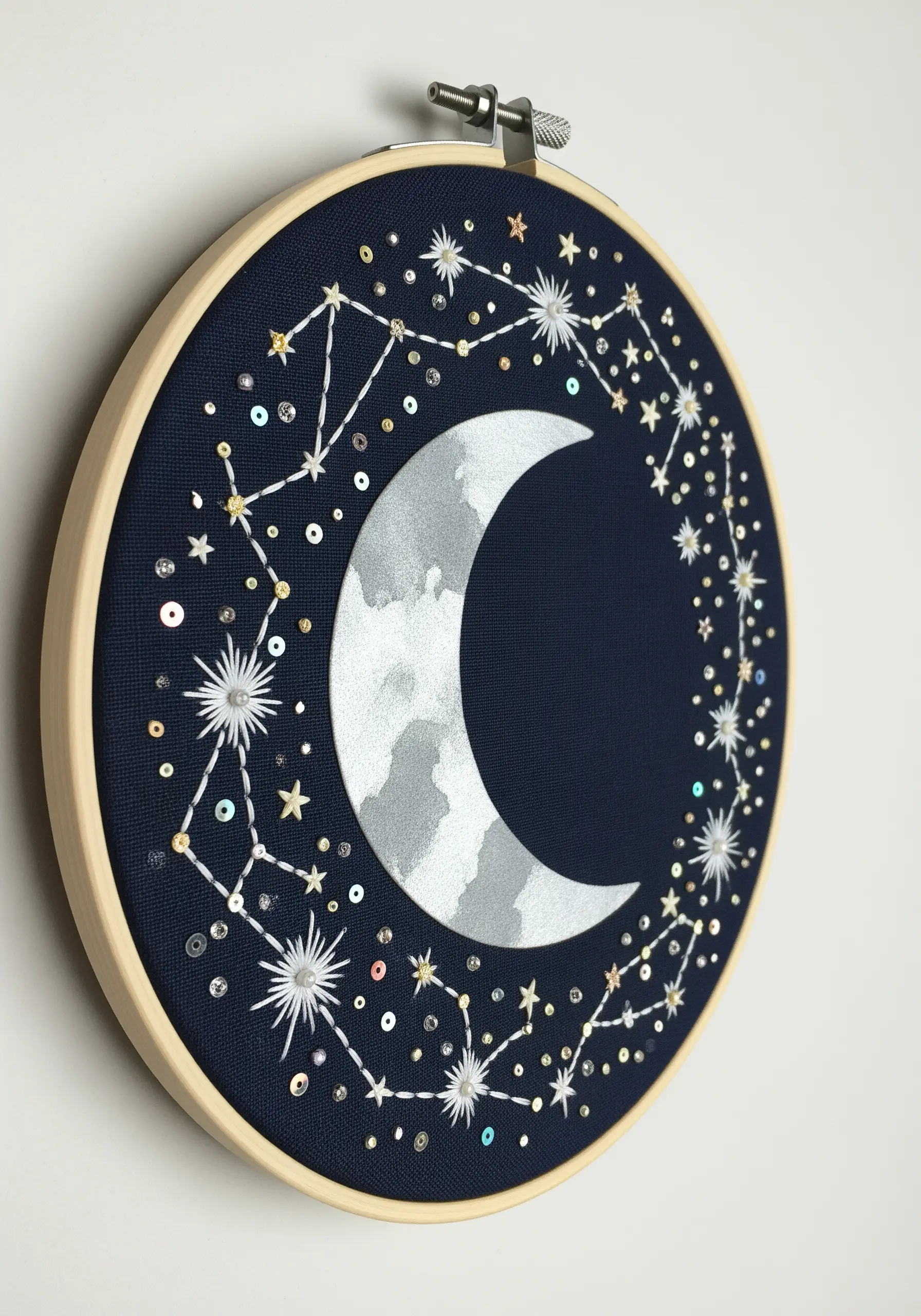 Celestial hoop art with a painted silver moon, sequin details, and embroidered star constellations.