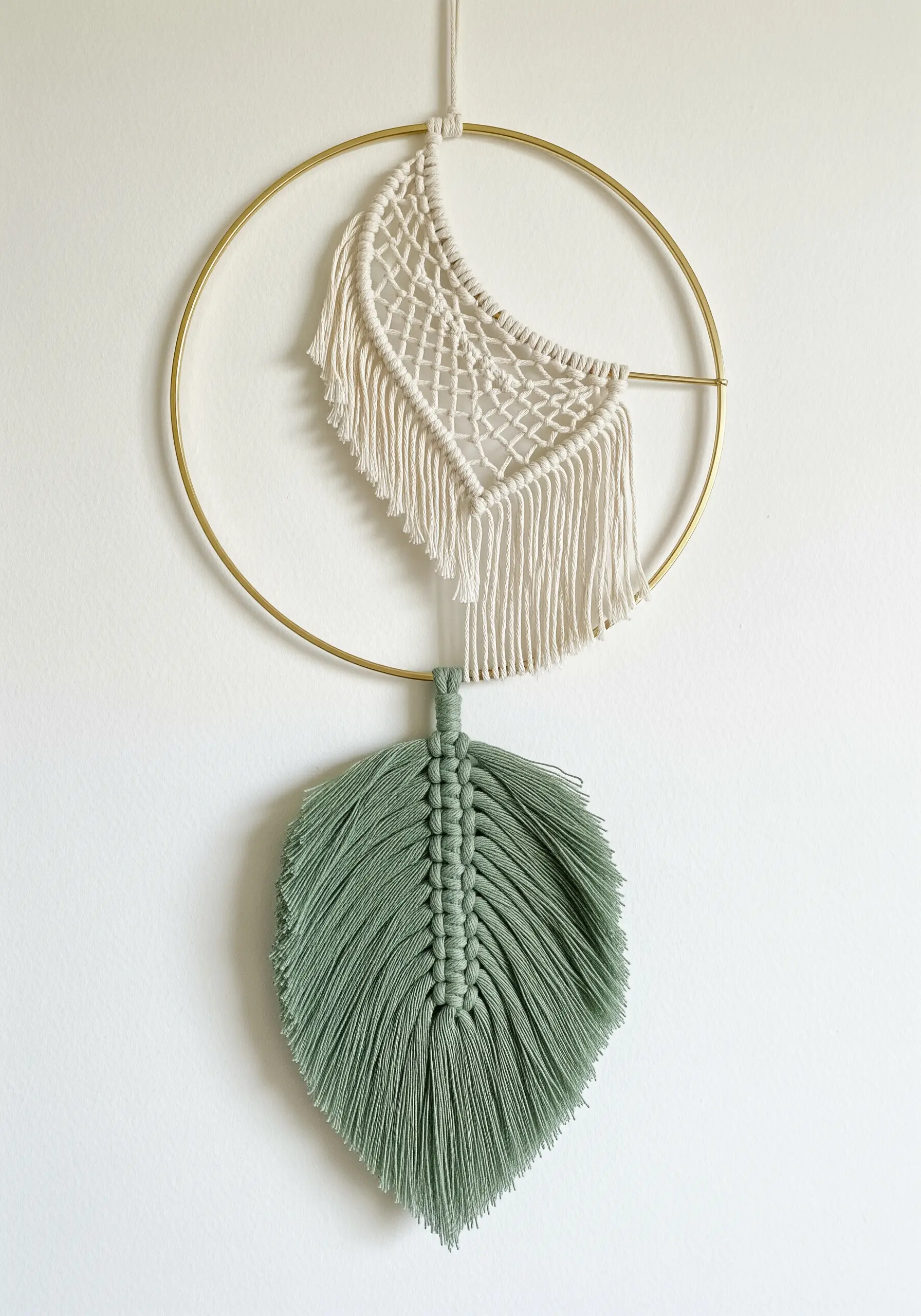 A minimalist wall hanging with a green macrame leaf and a white crescent moon on a gold hoop.