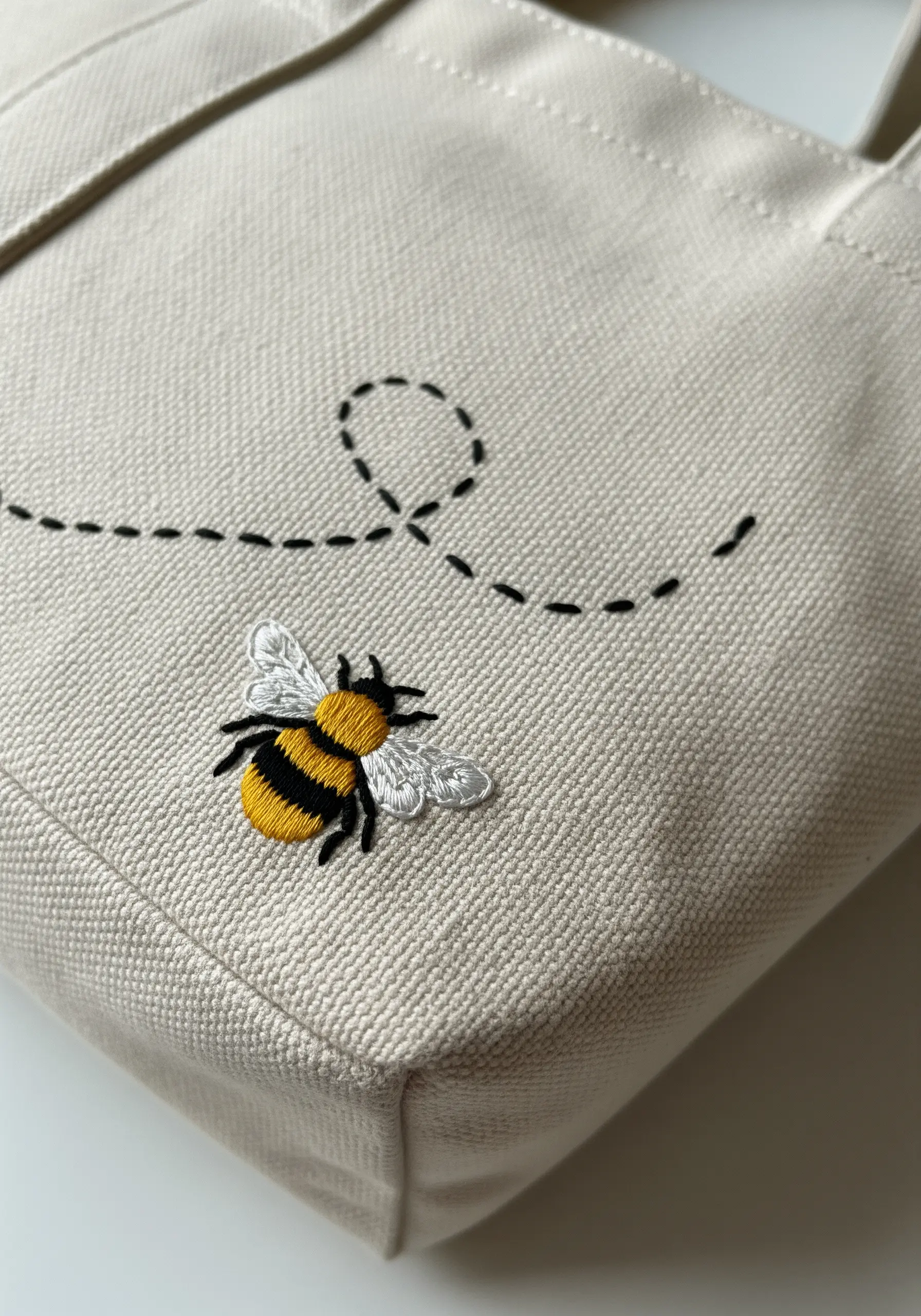 A small embroidered bee with satin stitch body and outlined wings on a canvas tote bag.