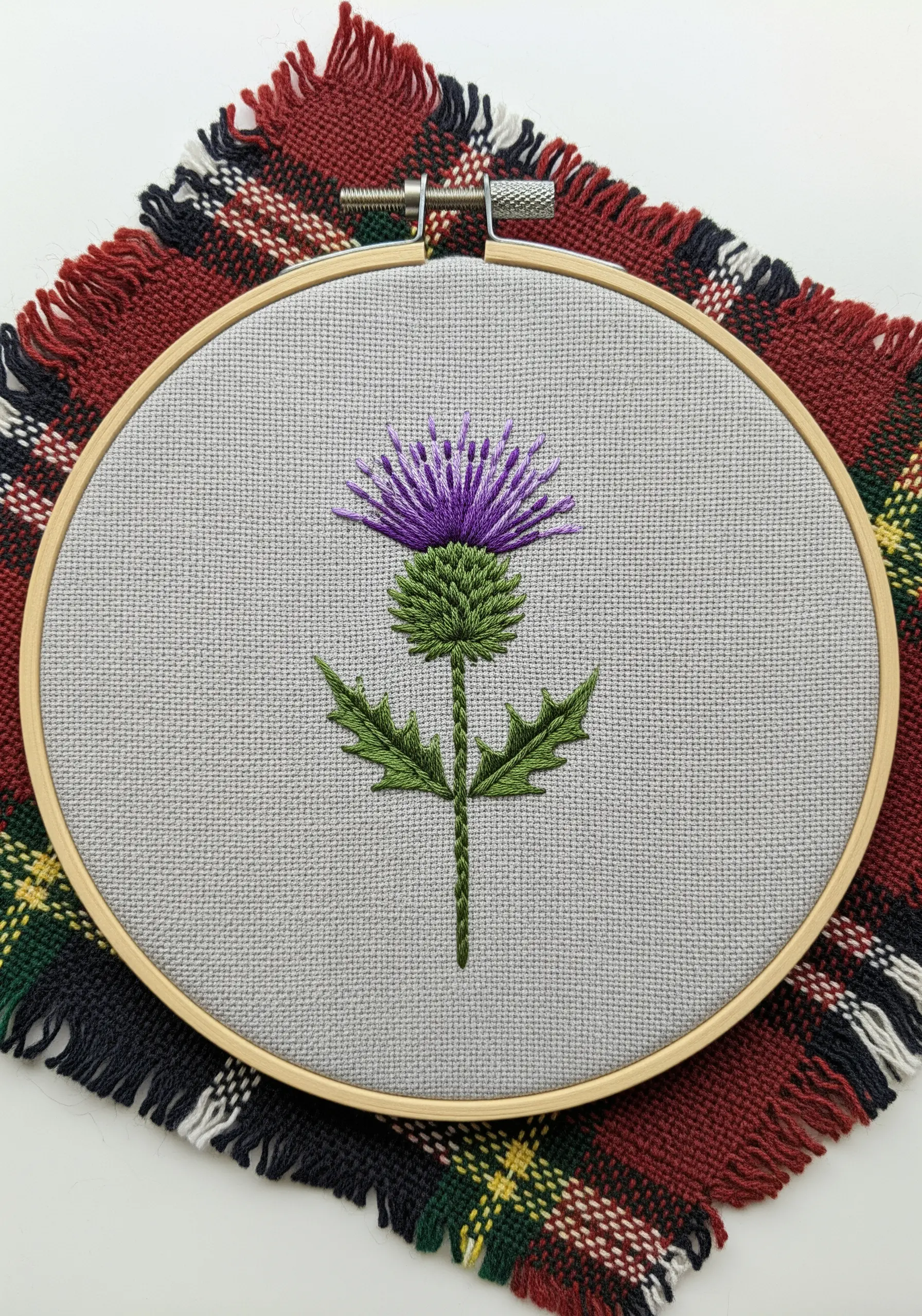 A Scottish thistle embroidered with purple and green thread on a gray fabric background.