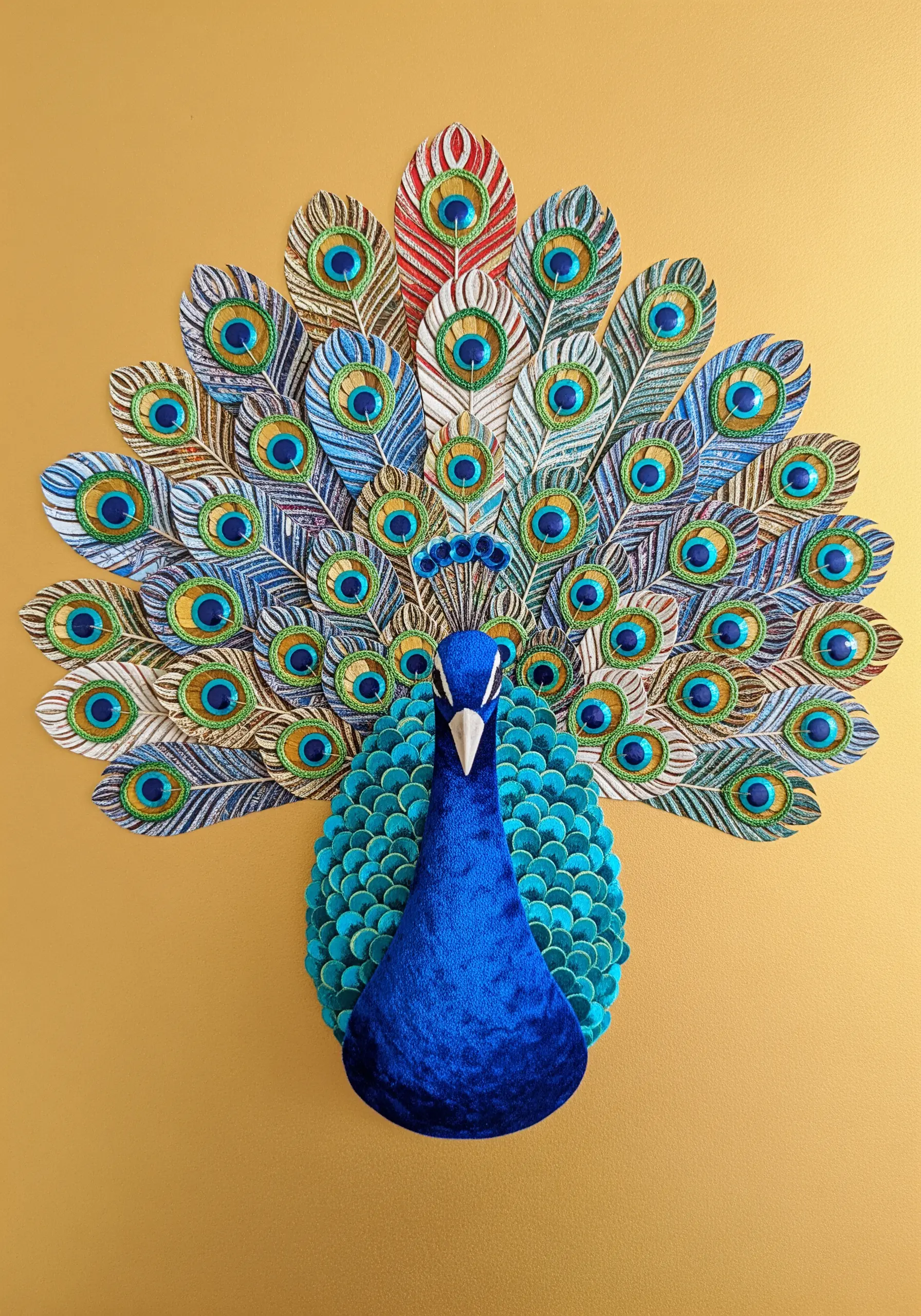 A peacock collage with a blue velvet body and layered paper tail feathers with beaded eyes.