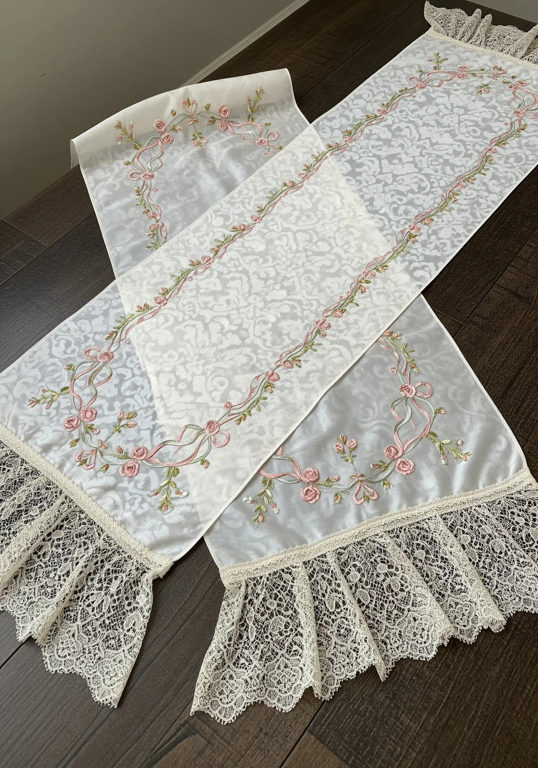 Embroidered pink roses and woven ribbons on a white damask table runner with ruffled lace.