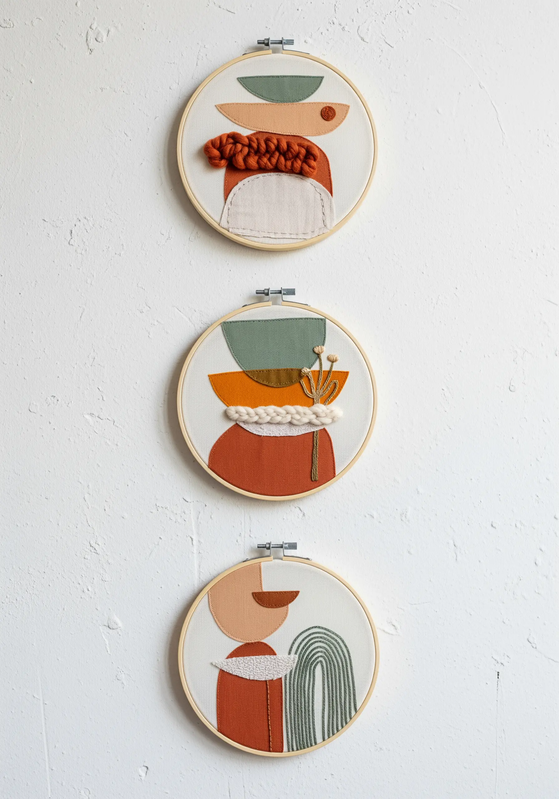 Three embroidery hoops with abstract collages of fabric and yarn in earth tones.