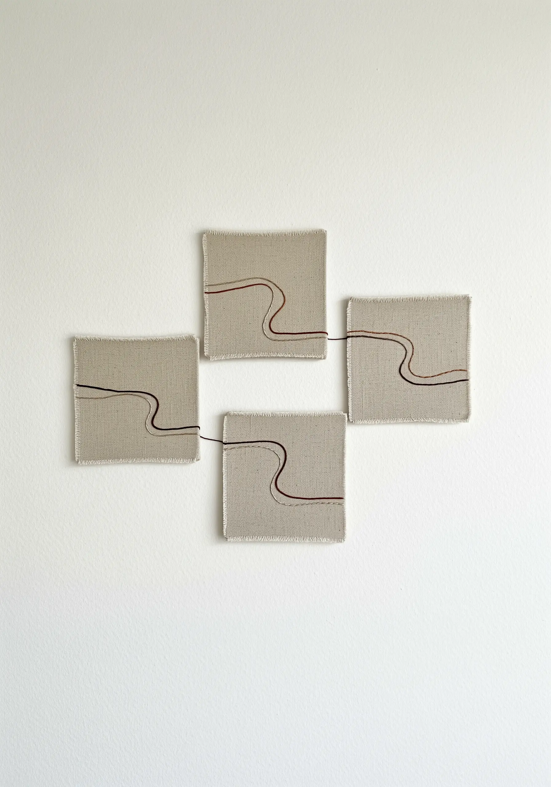 Four separate square linen canvases arranged on a wall, connected by a single, flowing embroidered line.