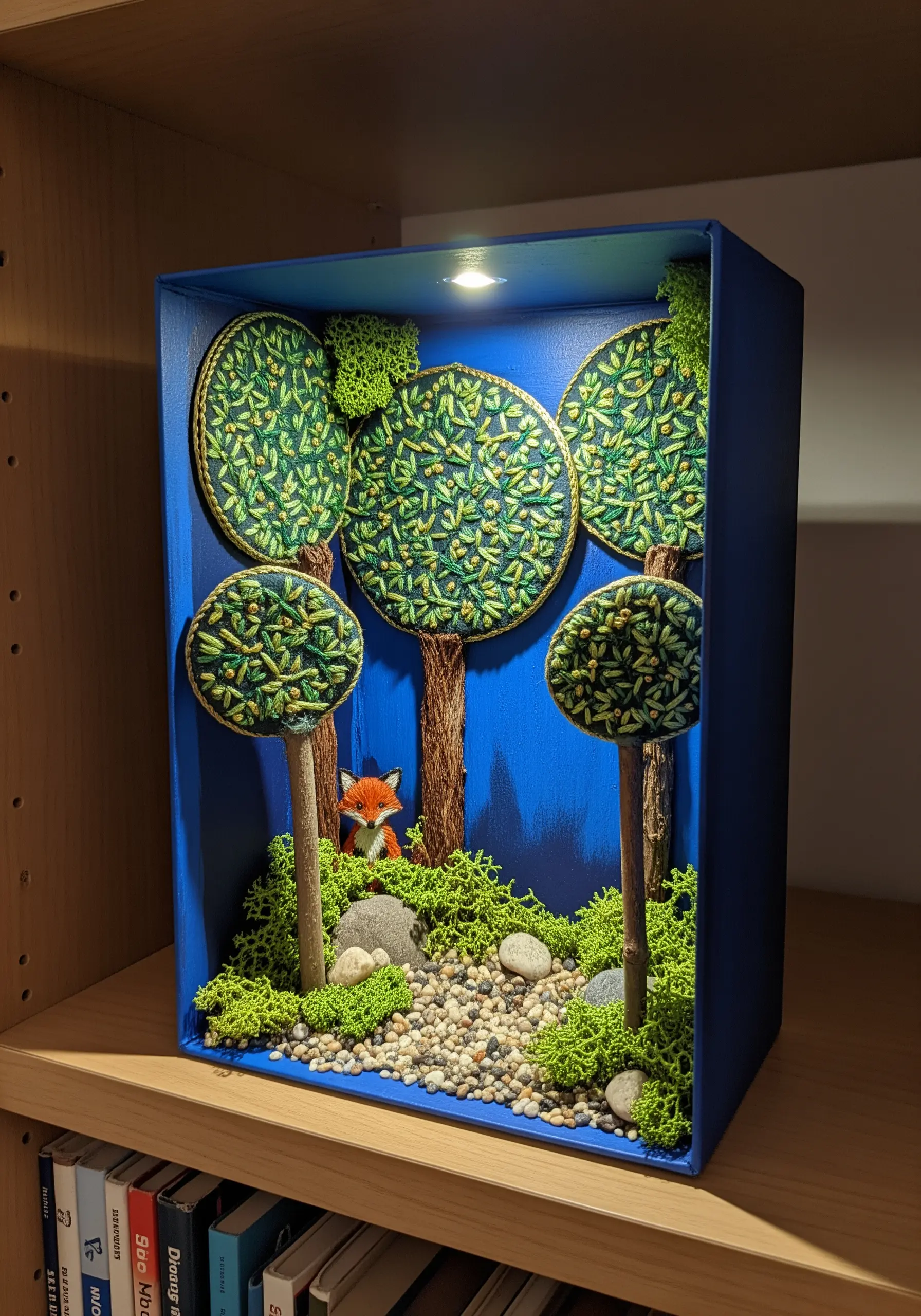 A diorama in a blue box featuring 3D embroidered trees, twigs, and a small fox figure.