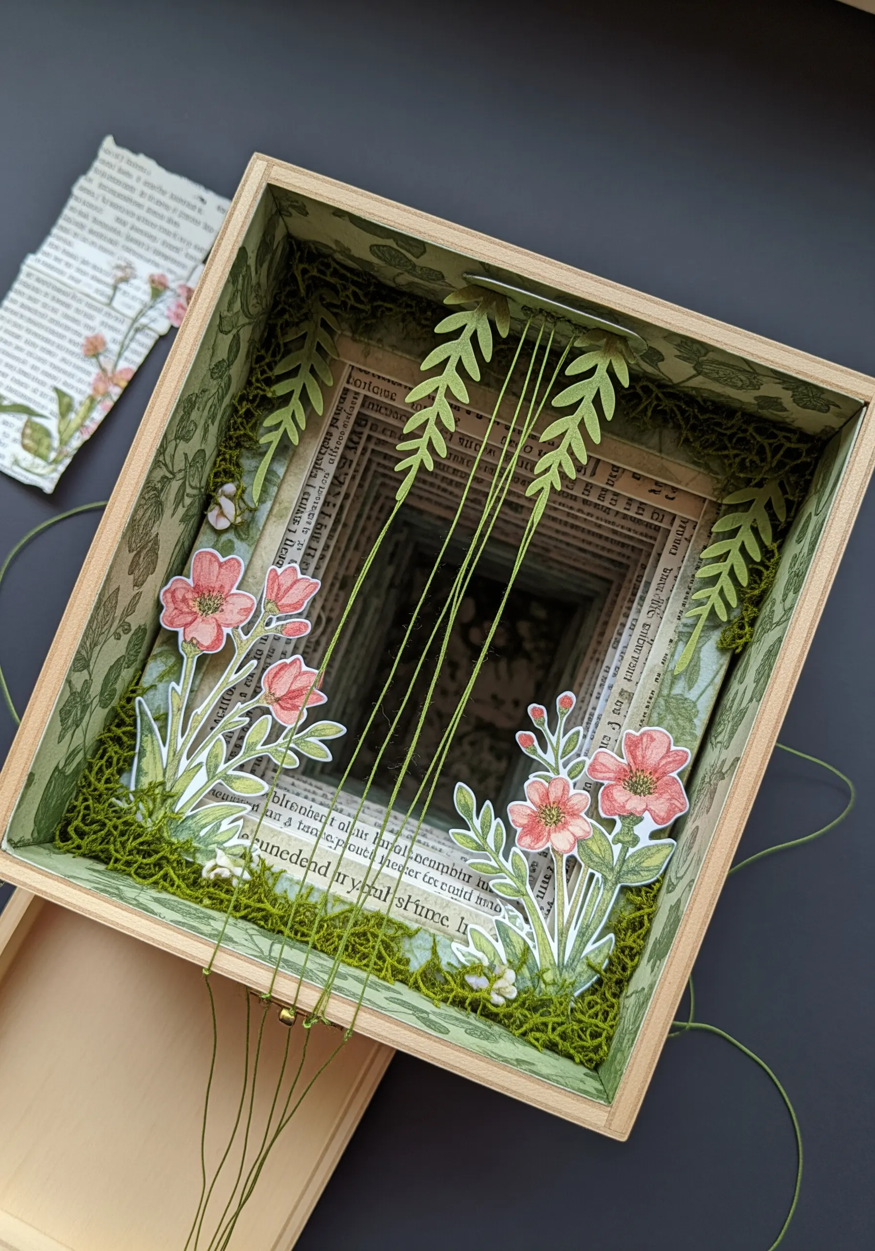 A 3D shadow box filled with layered paper botanicals, with green threads stretched vertically as vines.