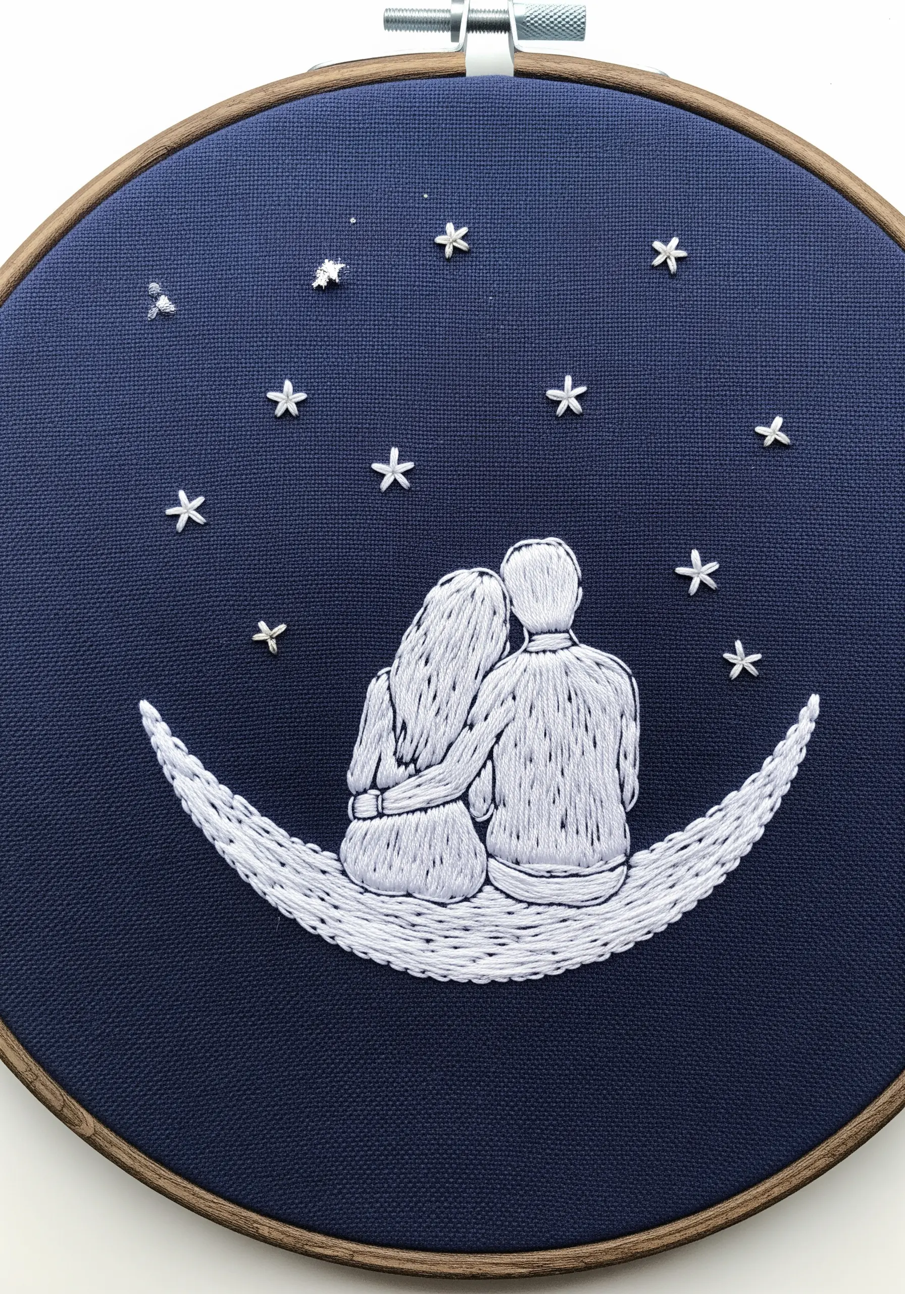 Embroidery of a couple sitting on a crescent moon against a navy background with white stars.