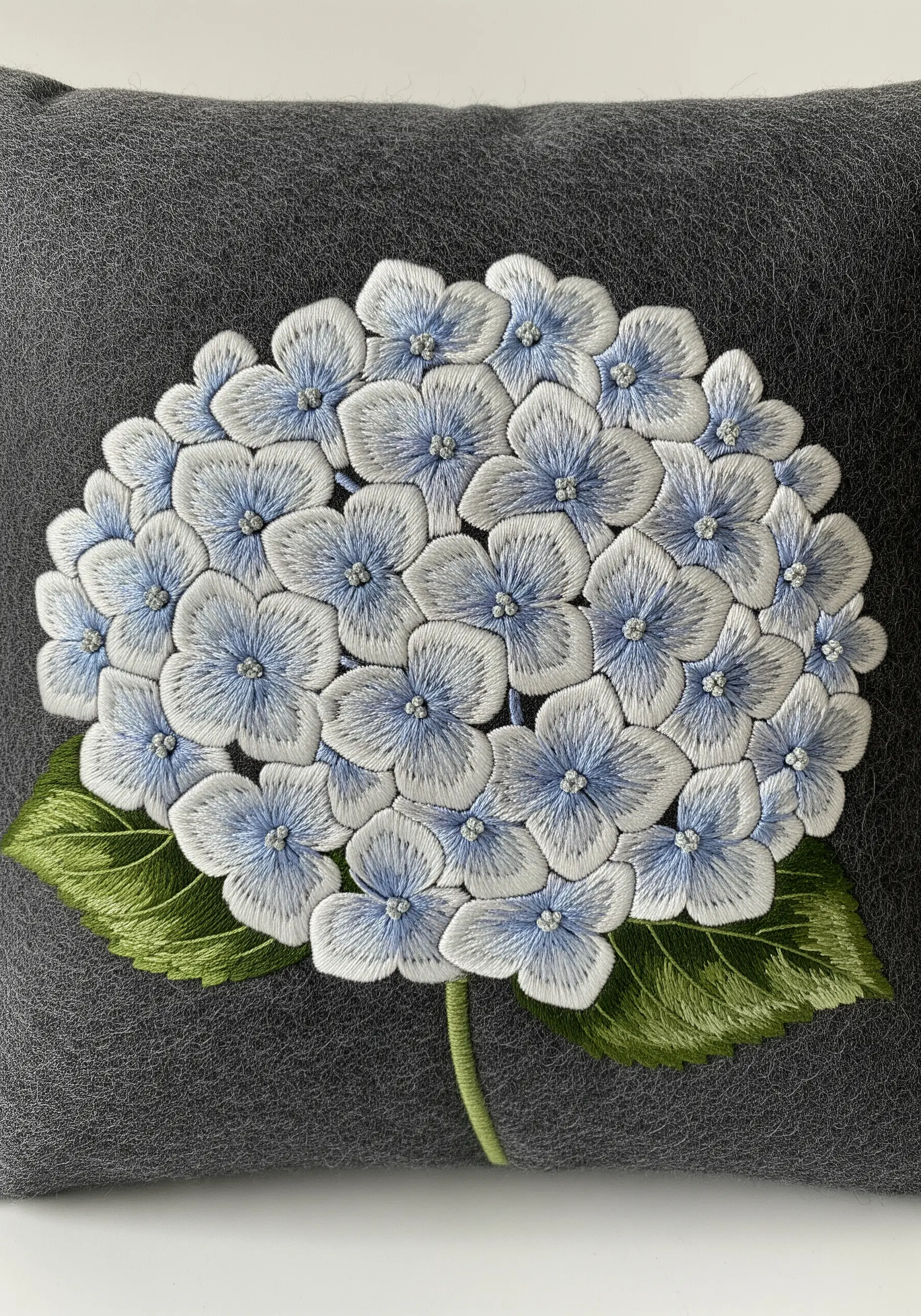 White and light blue hydrangea embroidered on a dark grey, textured wool pillow.