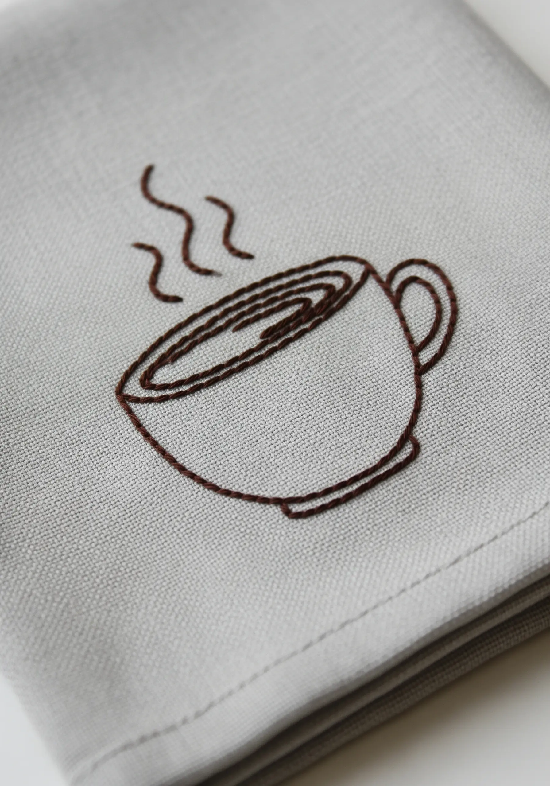 Minimalist line art embroidery of a steaming coffee cup on a grey linen napkin.