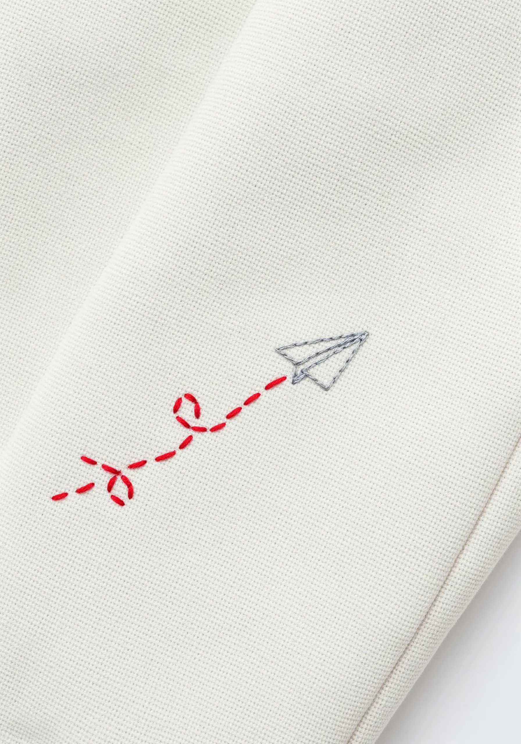Minimalist embroidery of a paper airplane with a red dotted-line trail on a white sleeve.