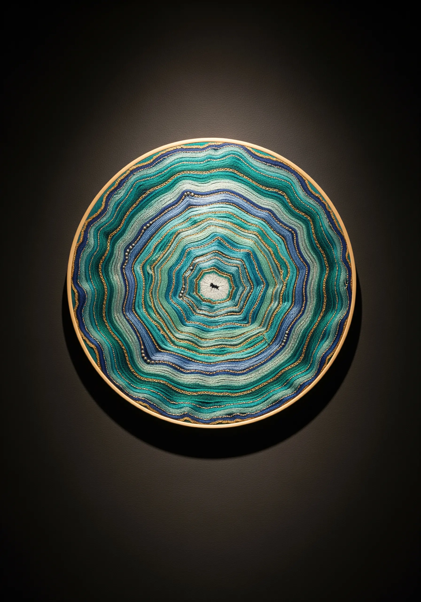 Circular embroidery hoop with concentric rings of blue, green, and gold thread resembling a geode.