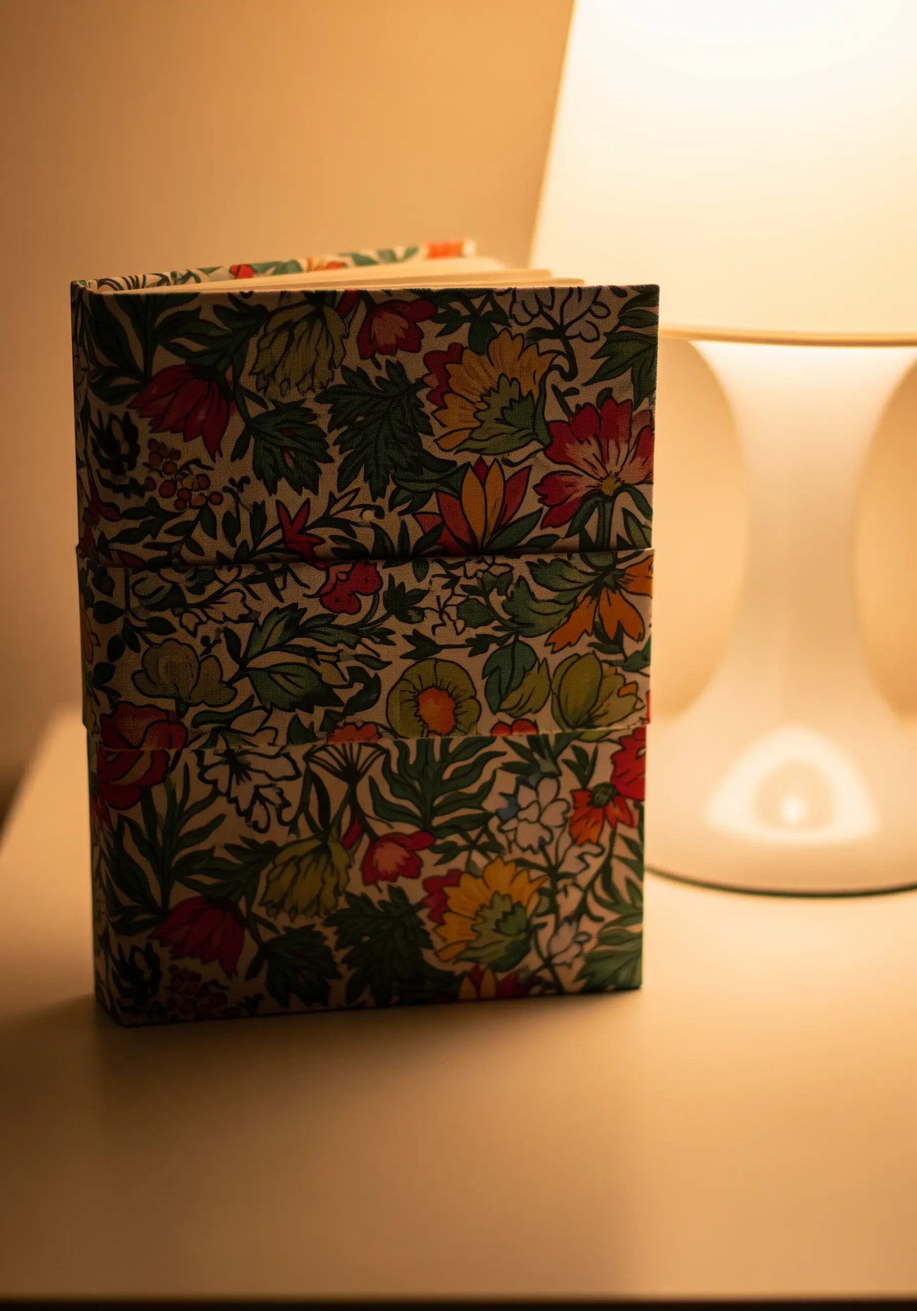 A notebook covered in a vibrant floral fabric, sitting on a desk next to a lamp.
