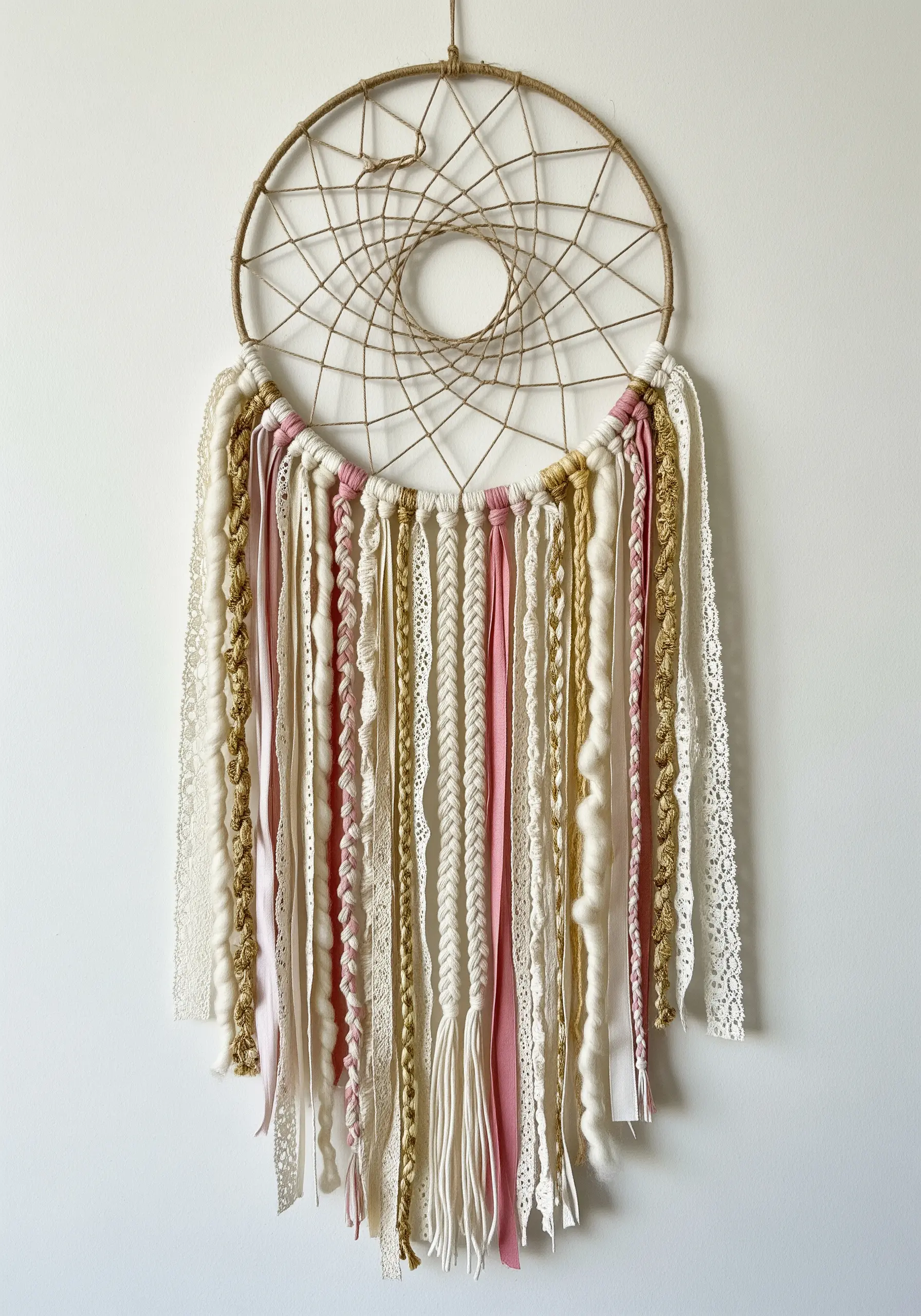 A dreamcatcher-style wall hanging with trailing ribbons, lace, fabric strips, and yarn in pink and cream.