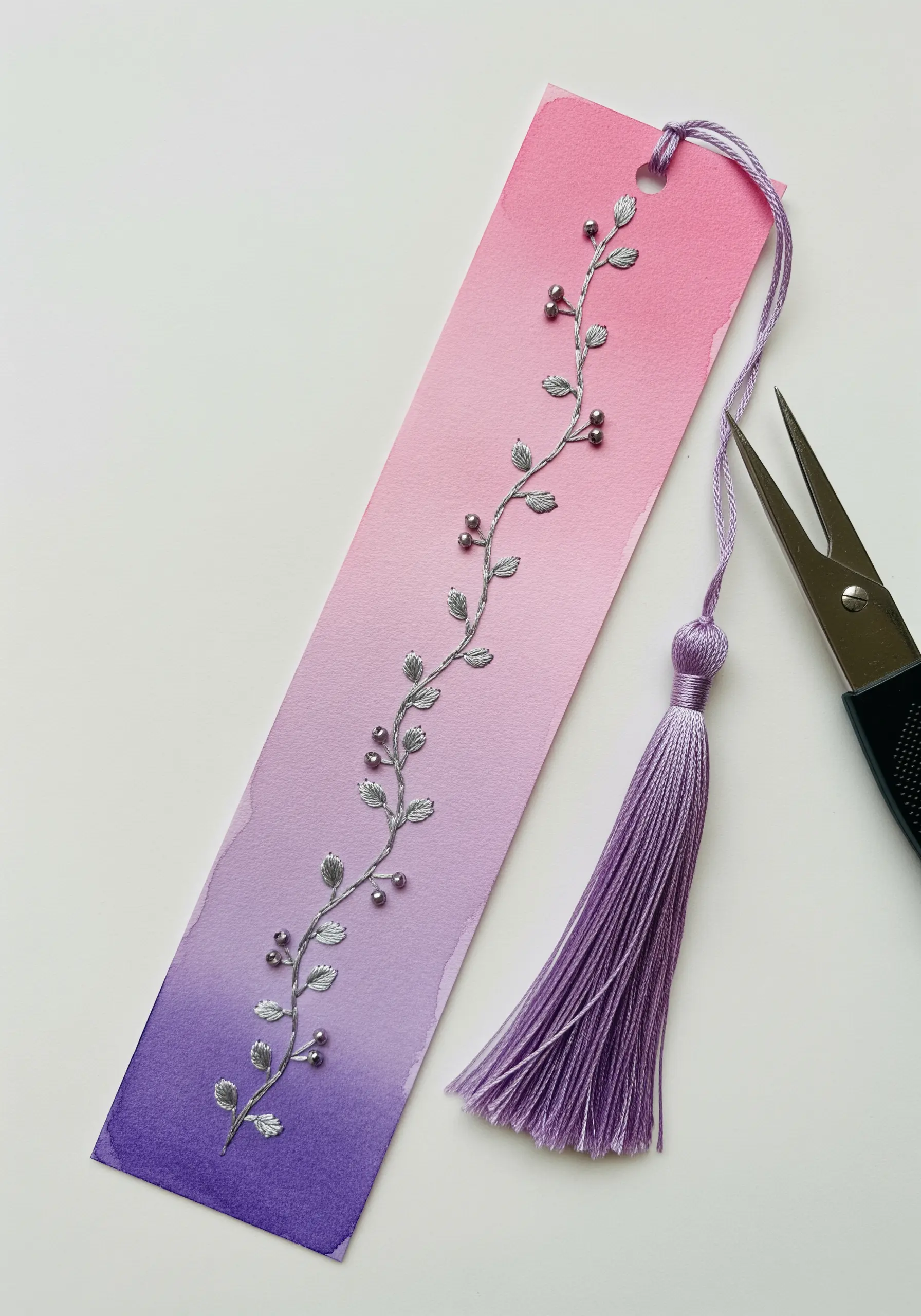 A watercolor gradient bookmark with a delicate vine embroidered in silver thread and beads.