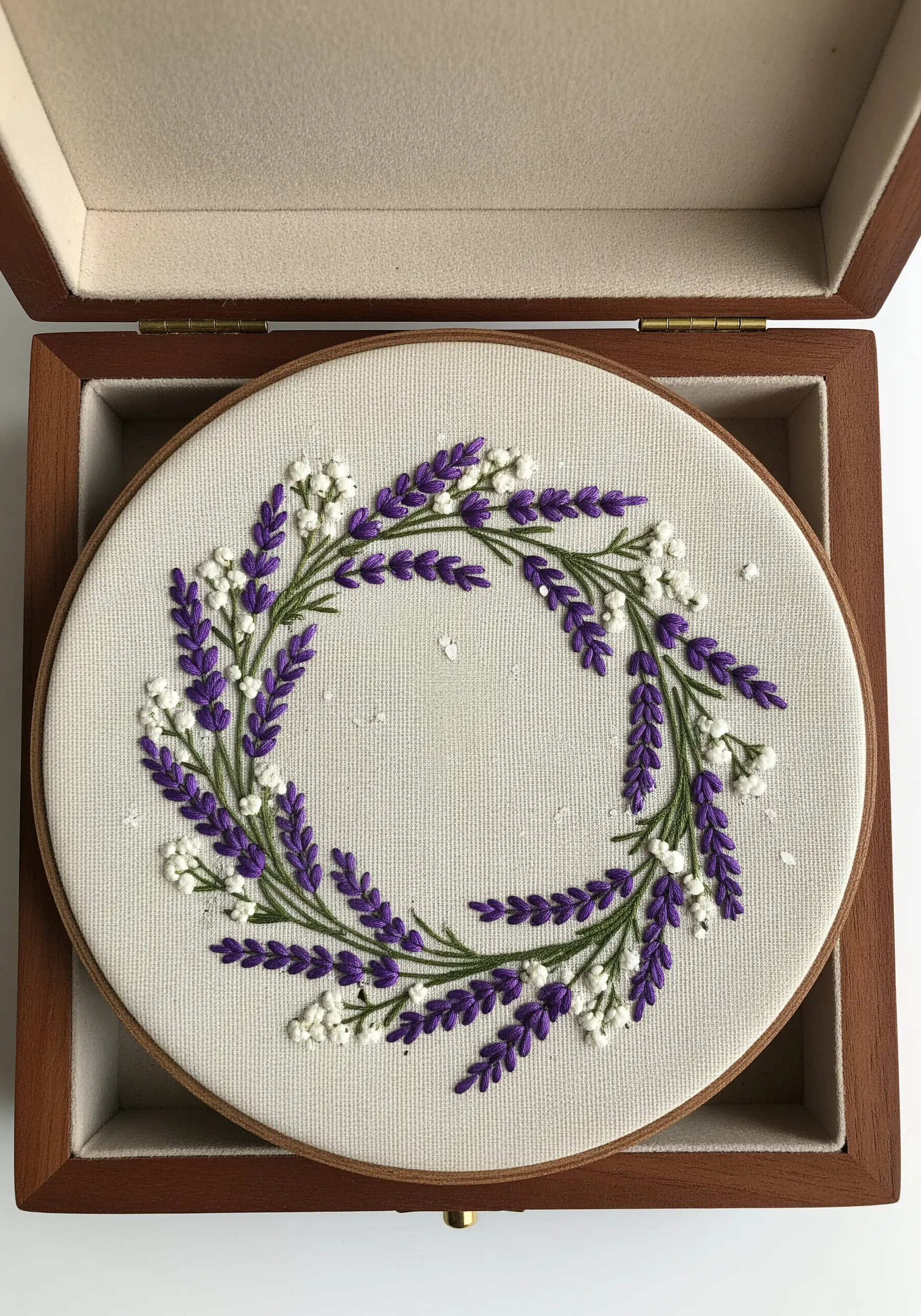 An embroidered wreath of lavender and baby's breath presented inside an open wooden box.
