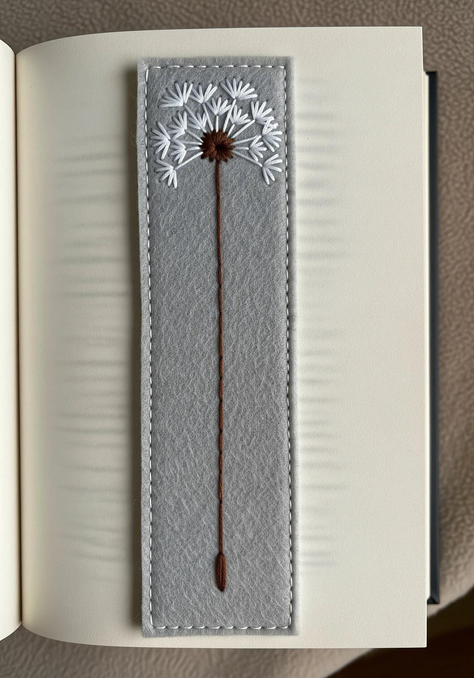An embroidered dandelion on a grey felt bookmark tucked inside a book.