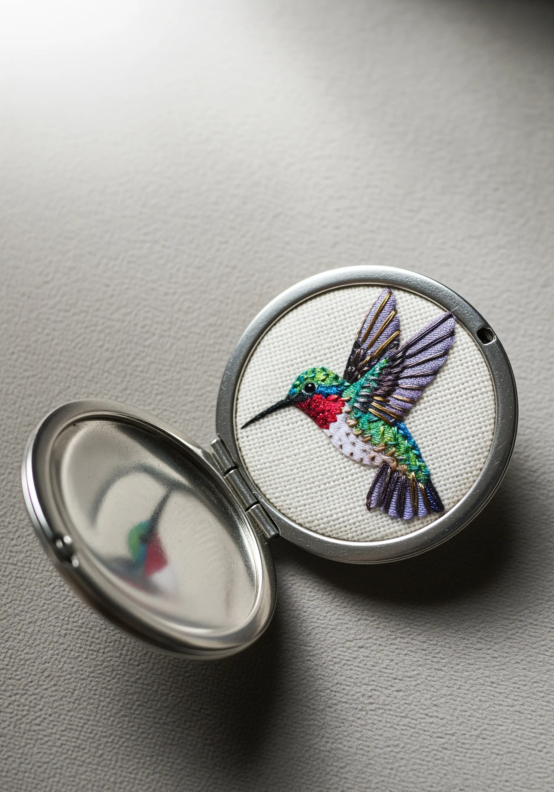 A tiny, detailed embroidered hummingbird made with metallic threads, set inside an open locket