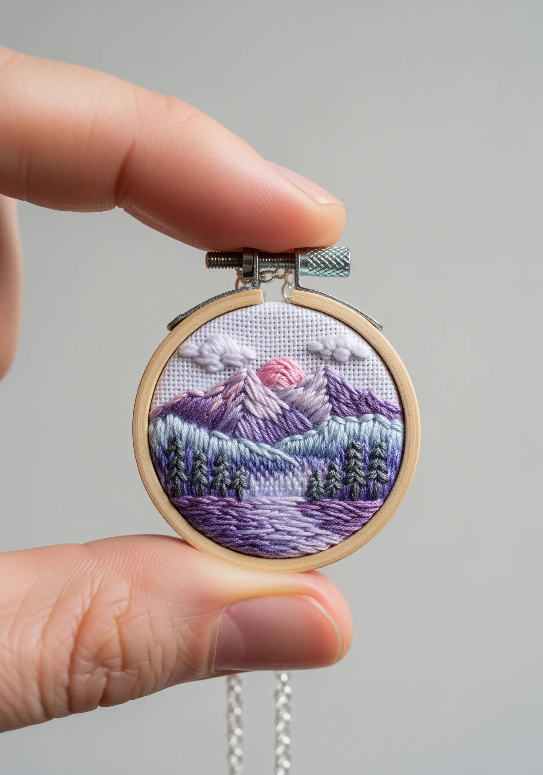 A tiny embroidery hoop pendant showing a mountain and forest landscape in purple hues.