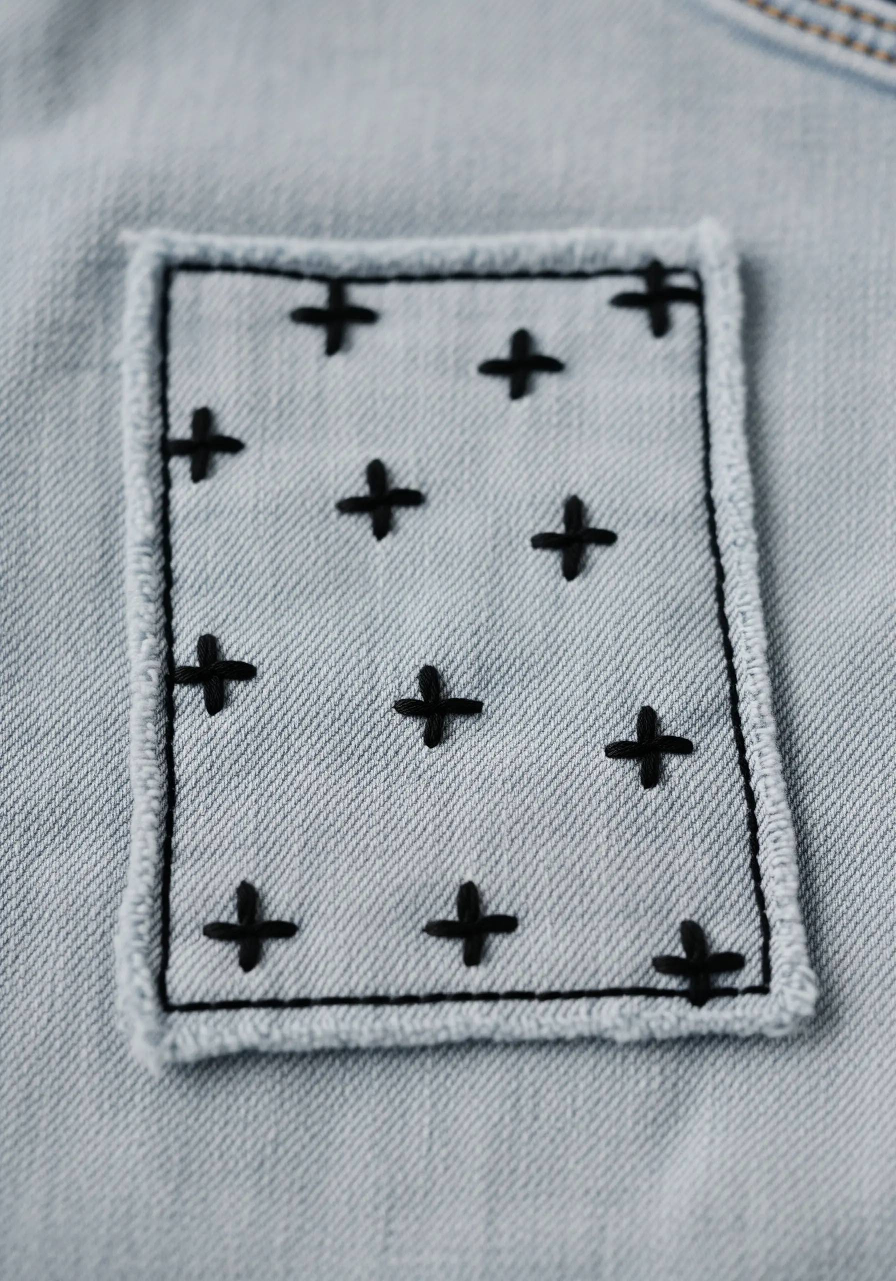 A light denim patch with black cross stitches and a raw edge, sewn onto a darker denim fabric.