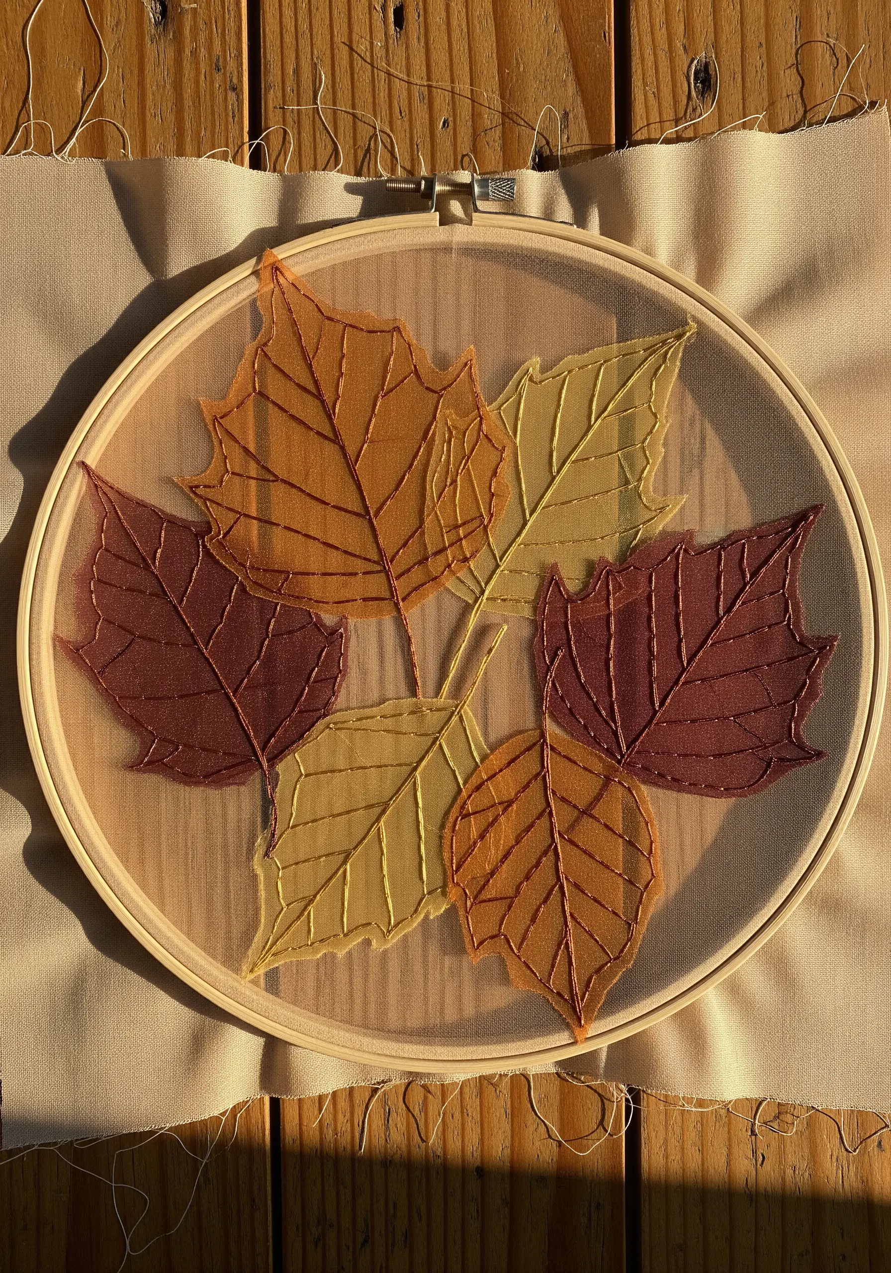 Sheer organza leaves in autumn colors stitched onto a transparent tulle base inside an embroidery hoop.