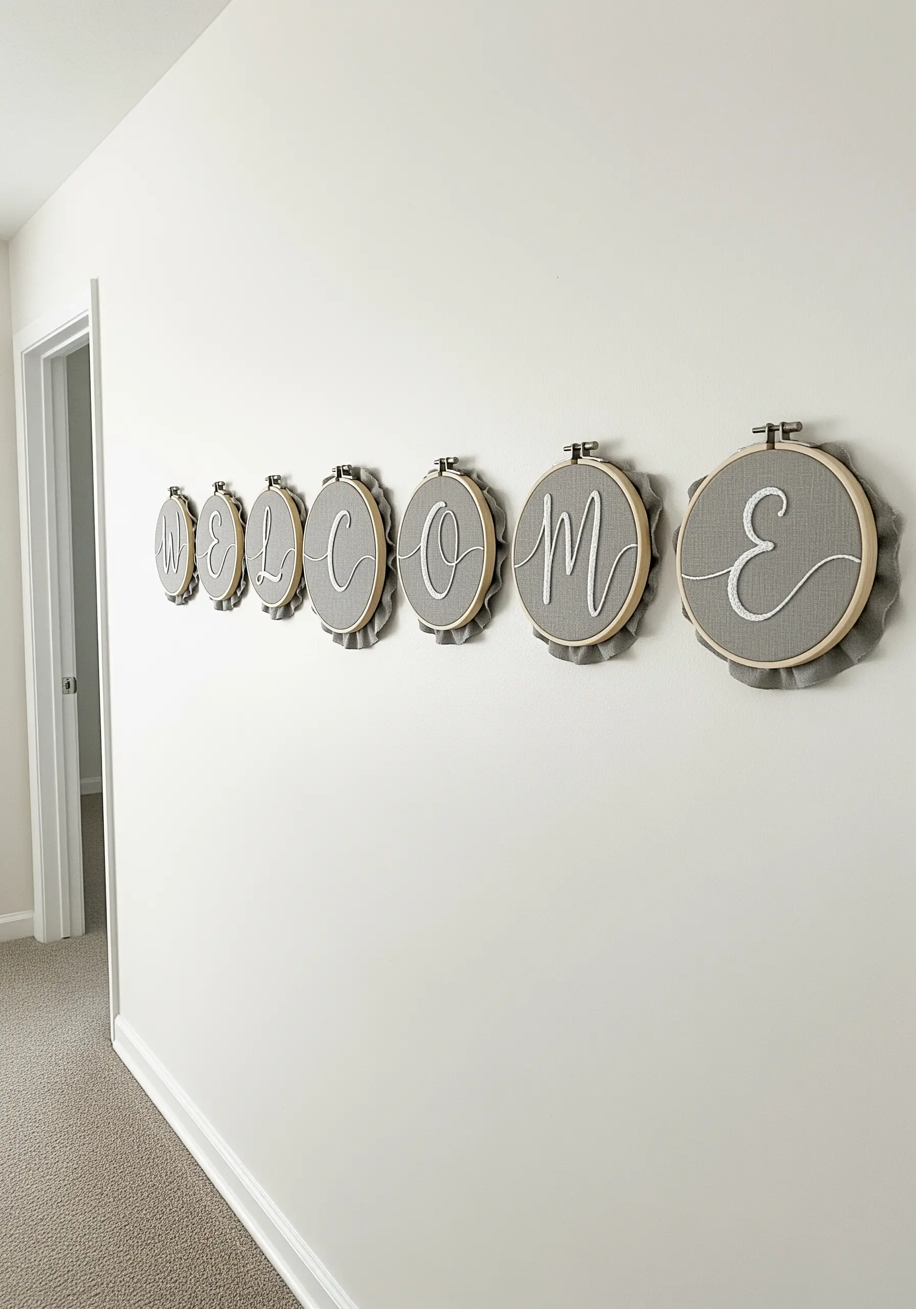 A series of small embroidery hoops spelling out 'Welcome' in elegant white script on grey fabric.