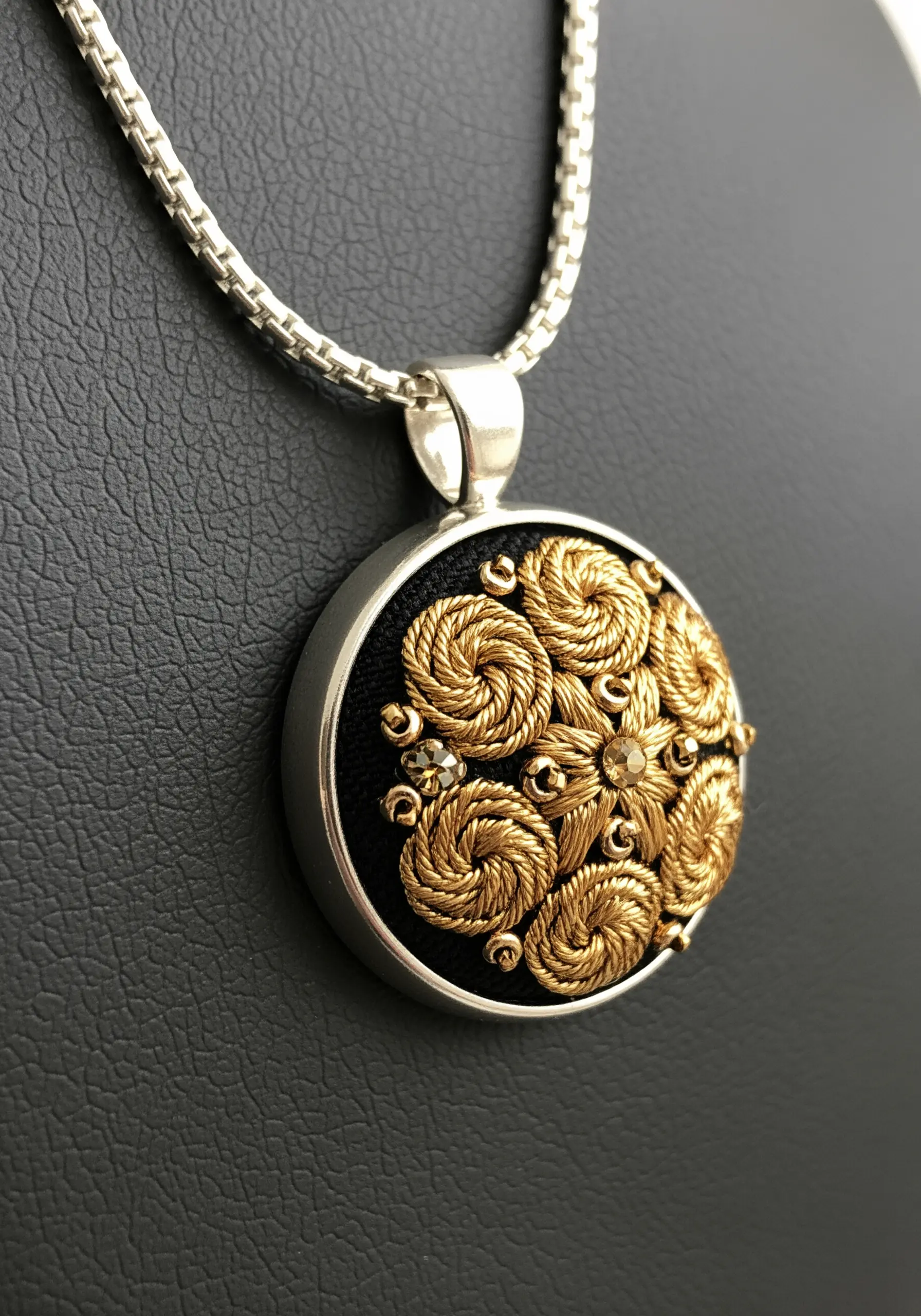 A close-up of a silver pendant filled with intricate gold metallic embroidery and tiny crystals.