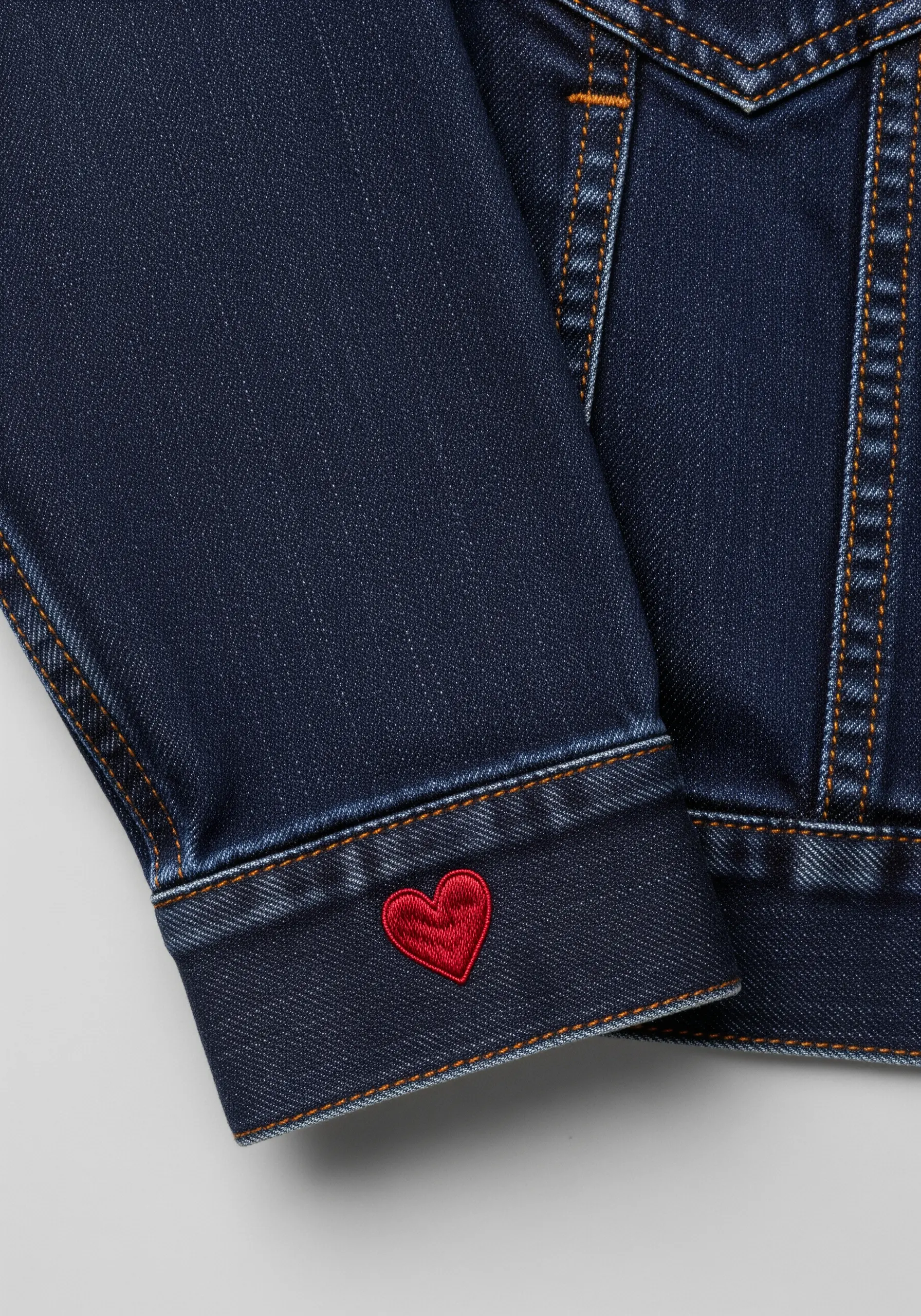 A small, vibrant red heart filled with satin stitch on the cuff of a dark denim jacket.