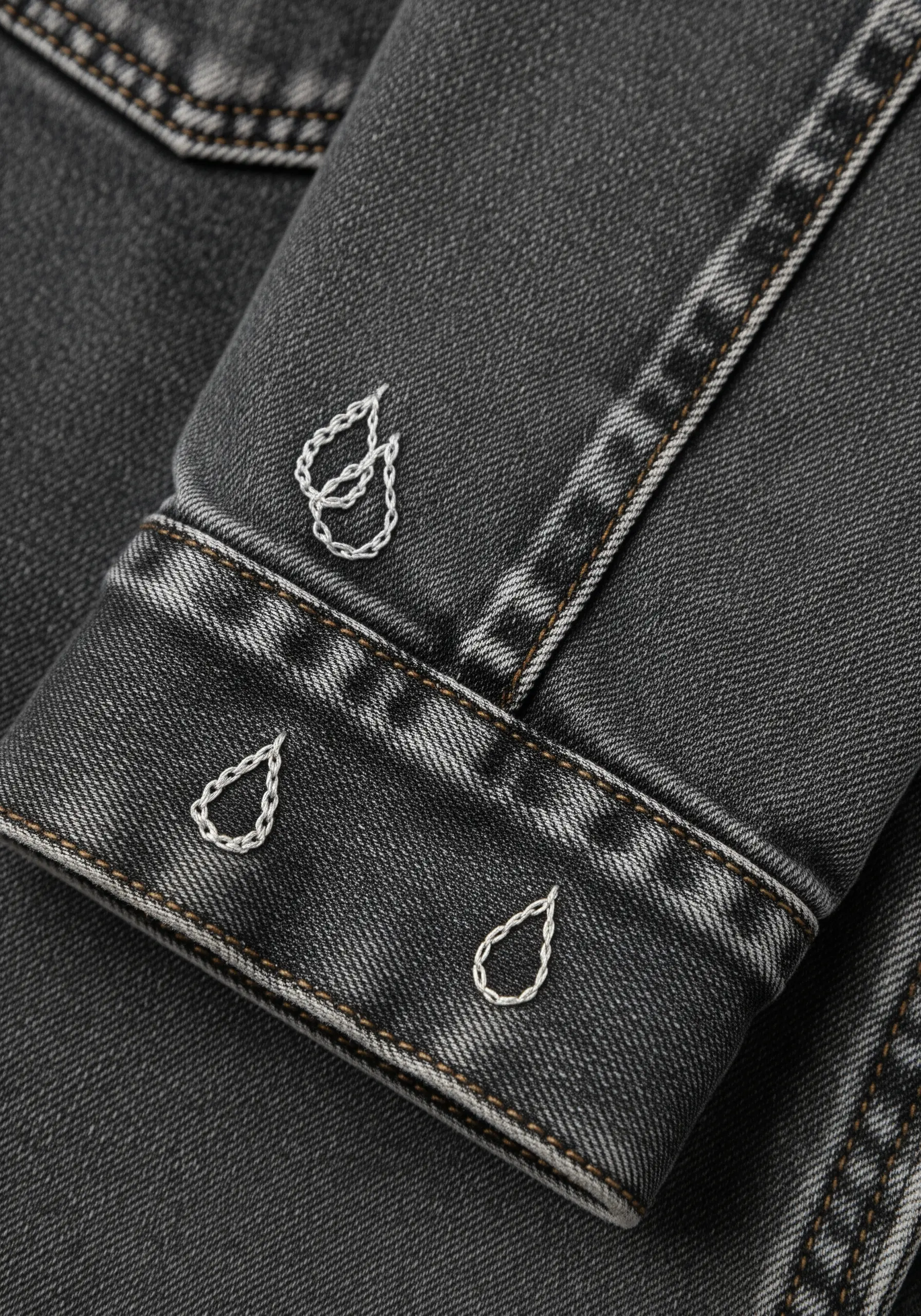 Small, simple teardrop outlines embroidered in white thread on a black denim jacket cuff.
