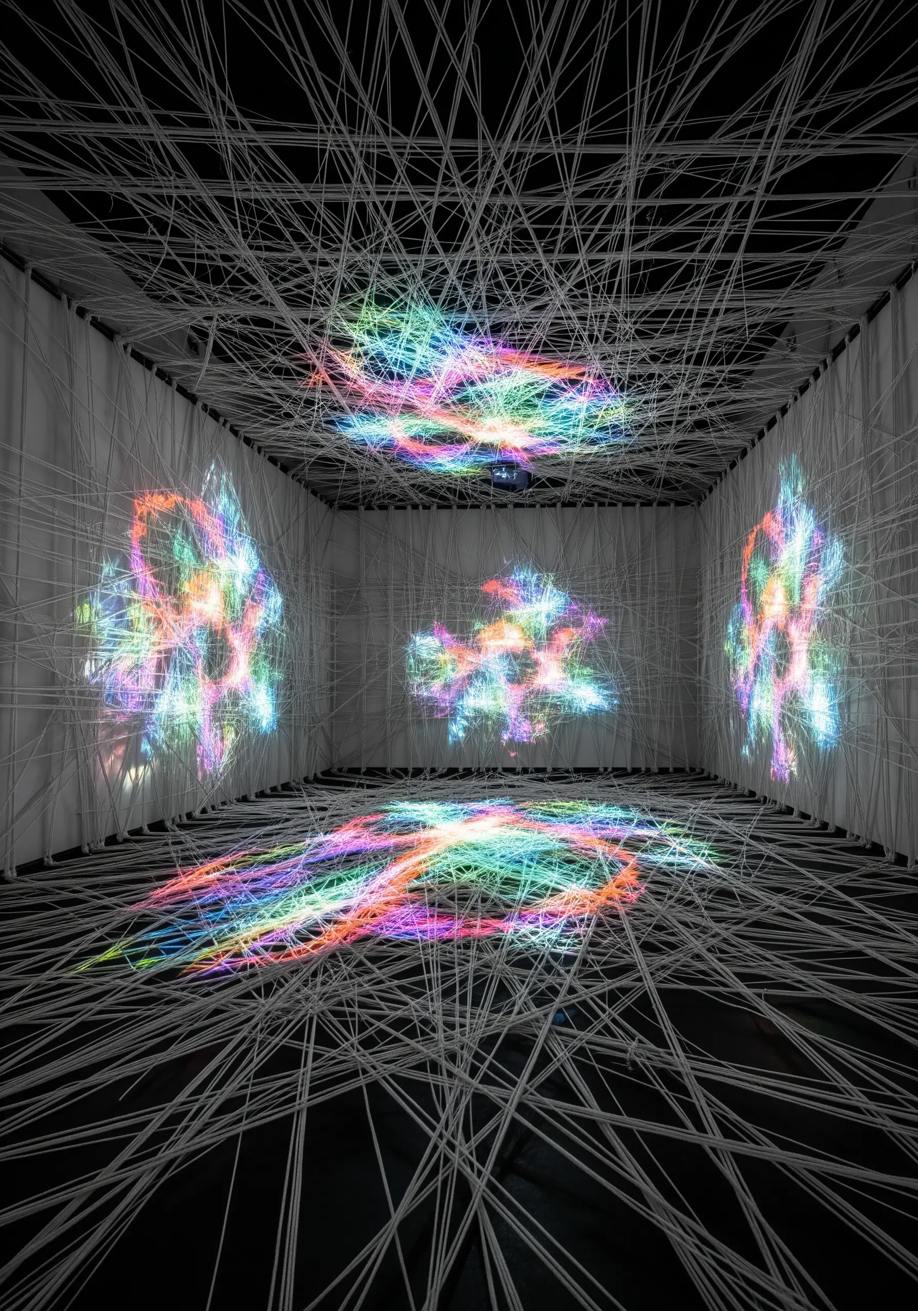 Colorful, abstract light patterns are projected onto a chaotic web of white string inside a dark room.