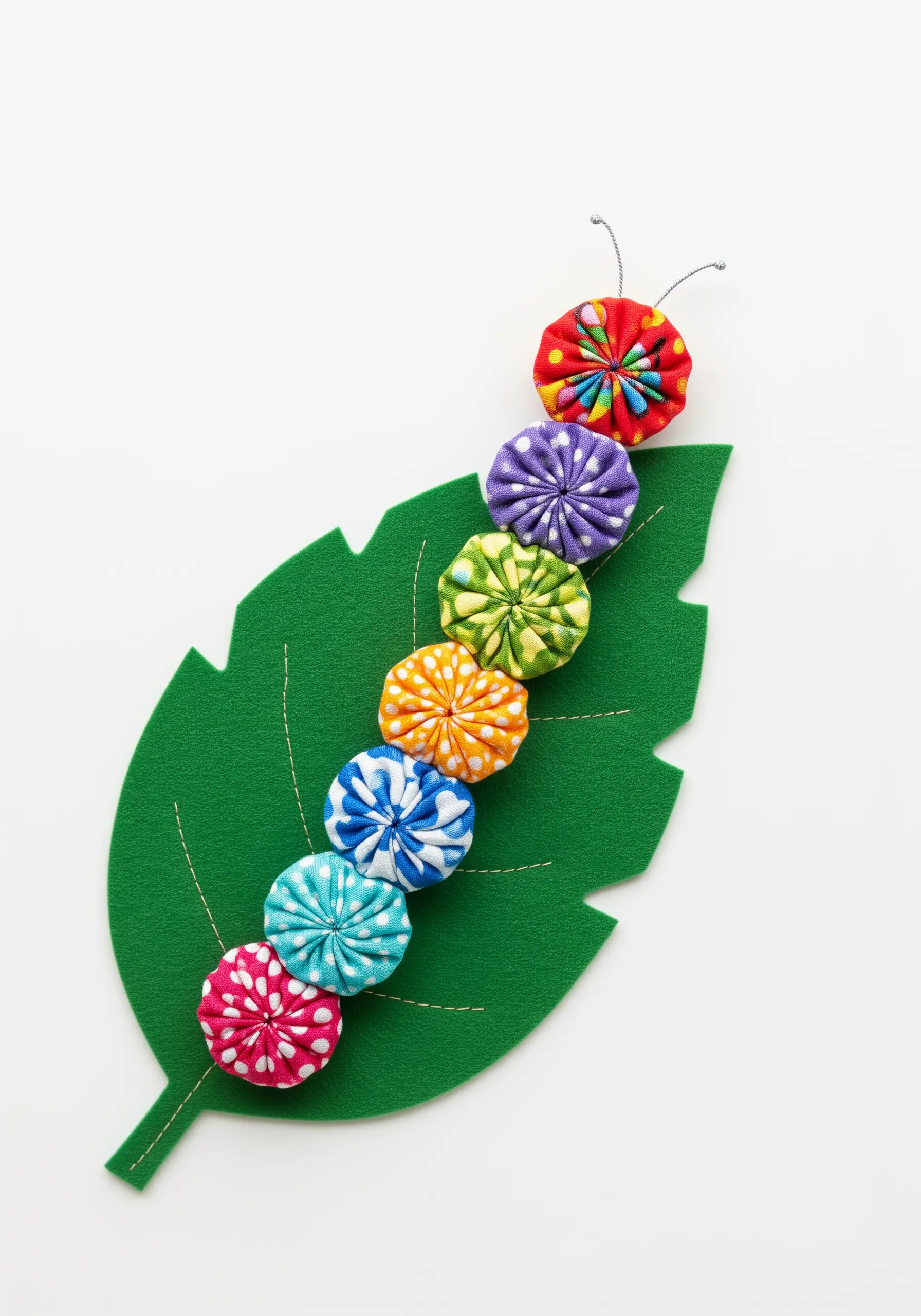 A colorful caterpillar made from fabric yo-yos sitting on a large green felt leaf.