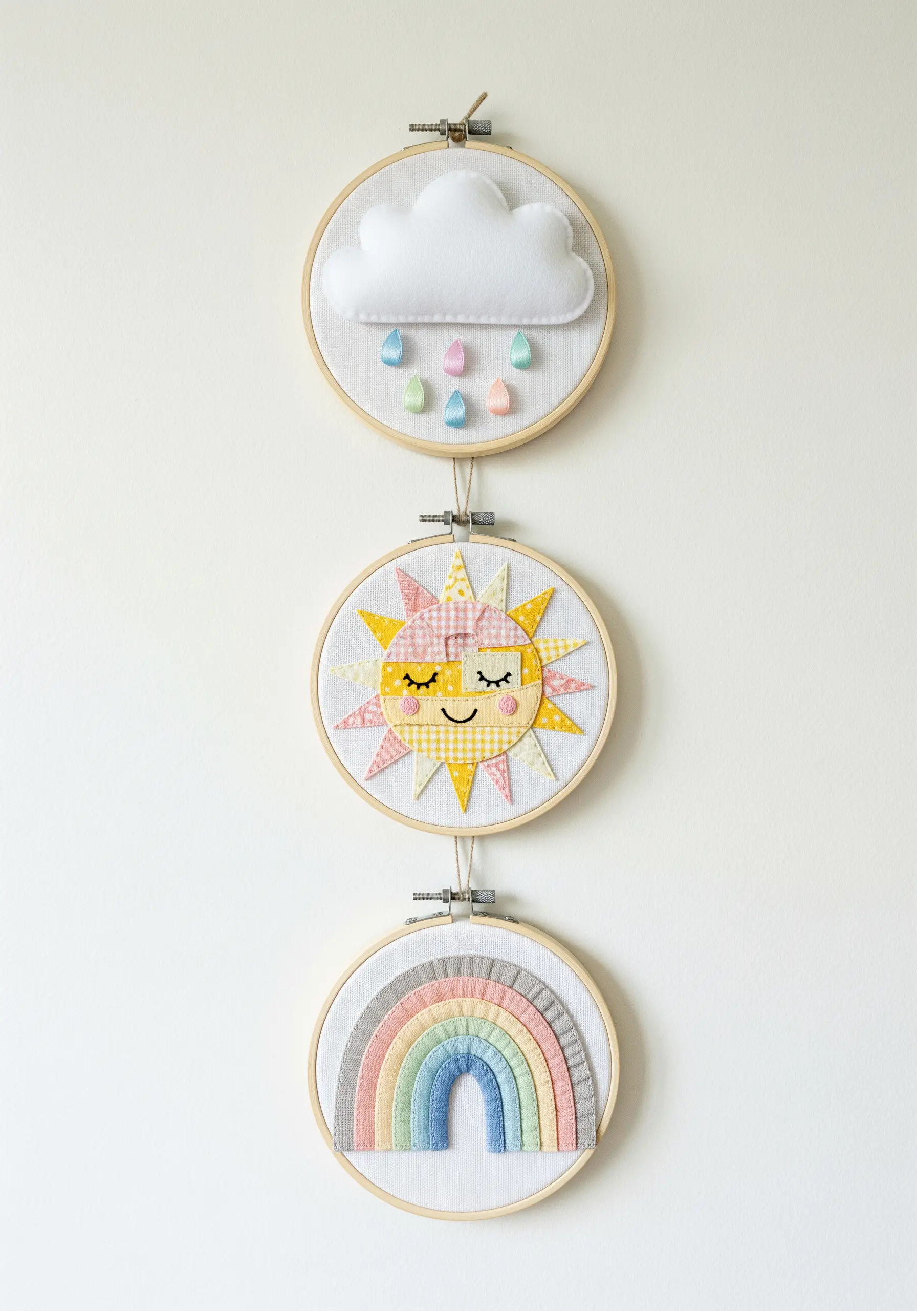 A trio of small embroidery hoops depicting a cloud, sun, and rainbow in pastel fabrics.
